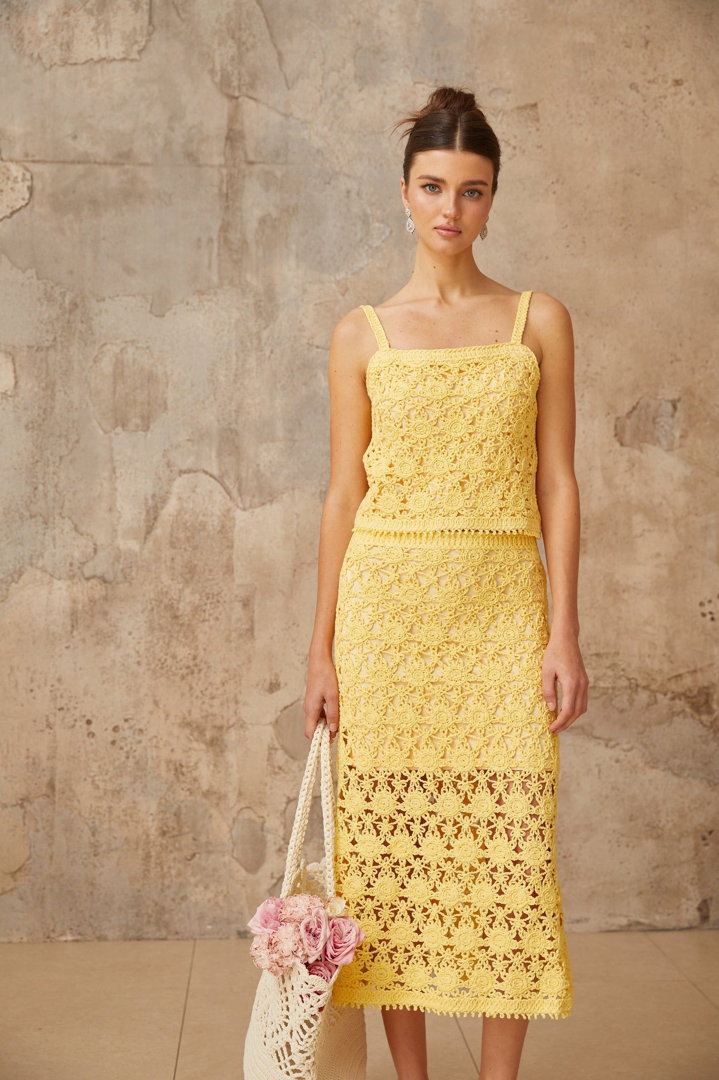 A young woman wearing a bright yellow crochet knit matching set consisting of a cropped top and mini skirt standing against a plain background