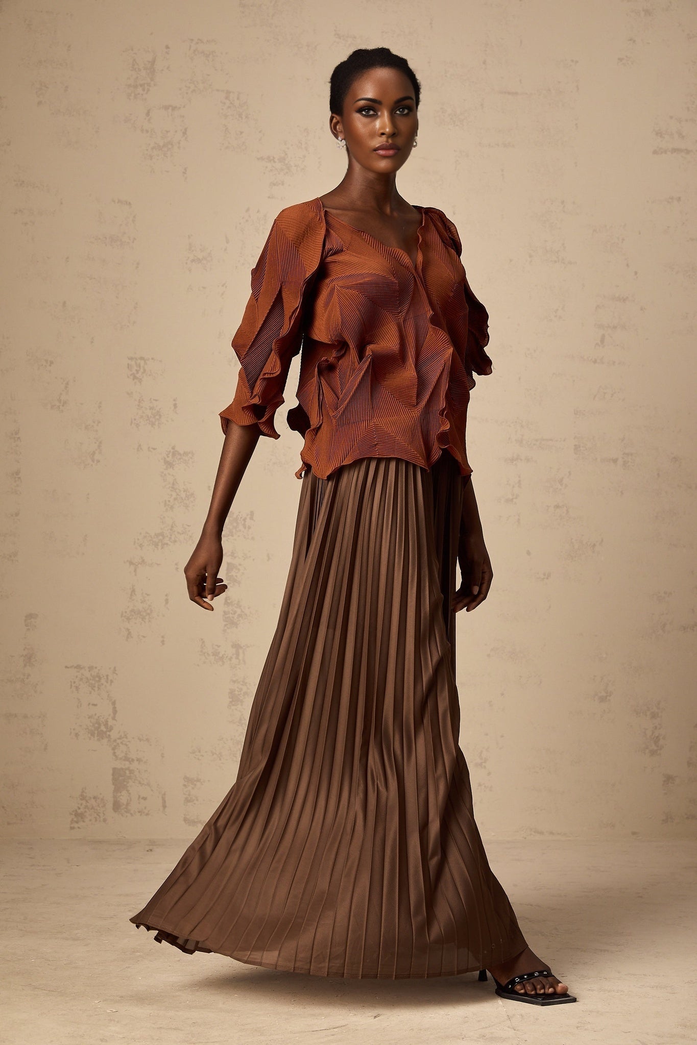 A young woman wearing a brown cropped top with crease details and a matching midi skirt standing against a plain background