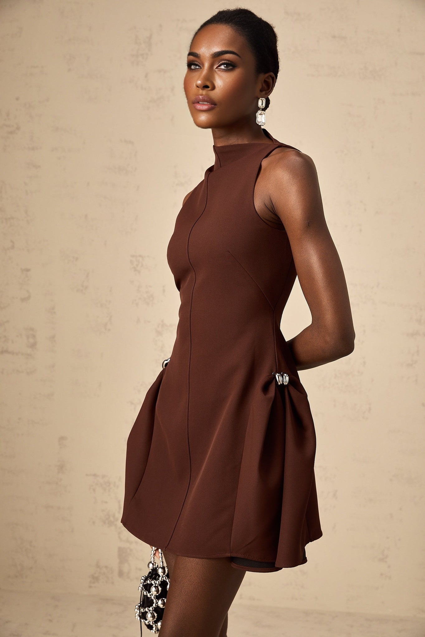 A young woman wearing a short brown dress with an asymmetric neckline and puffed sleeves standing against a plain background