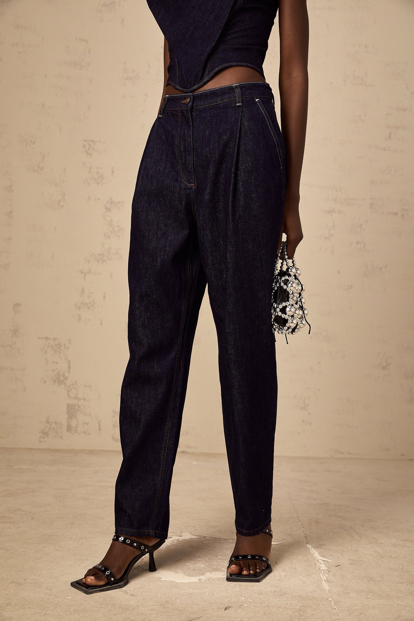 Blue high rise jeans with pleated details on the front worn by a model standing against a plain background