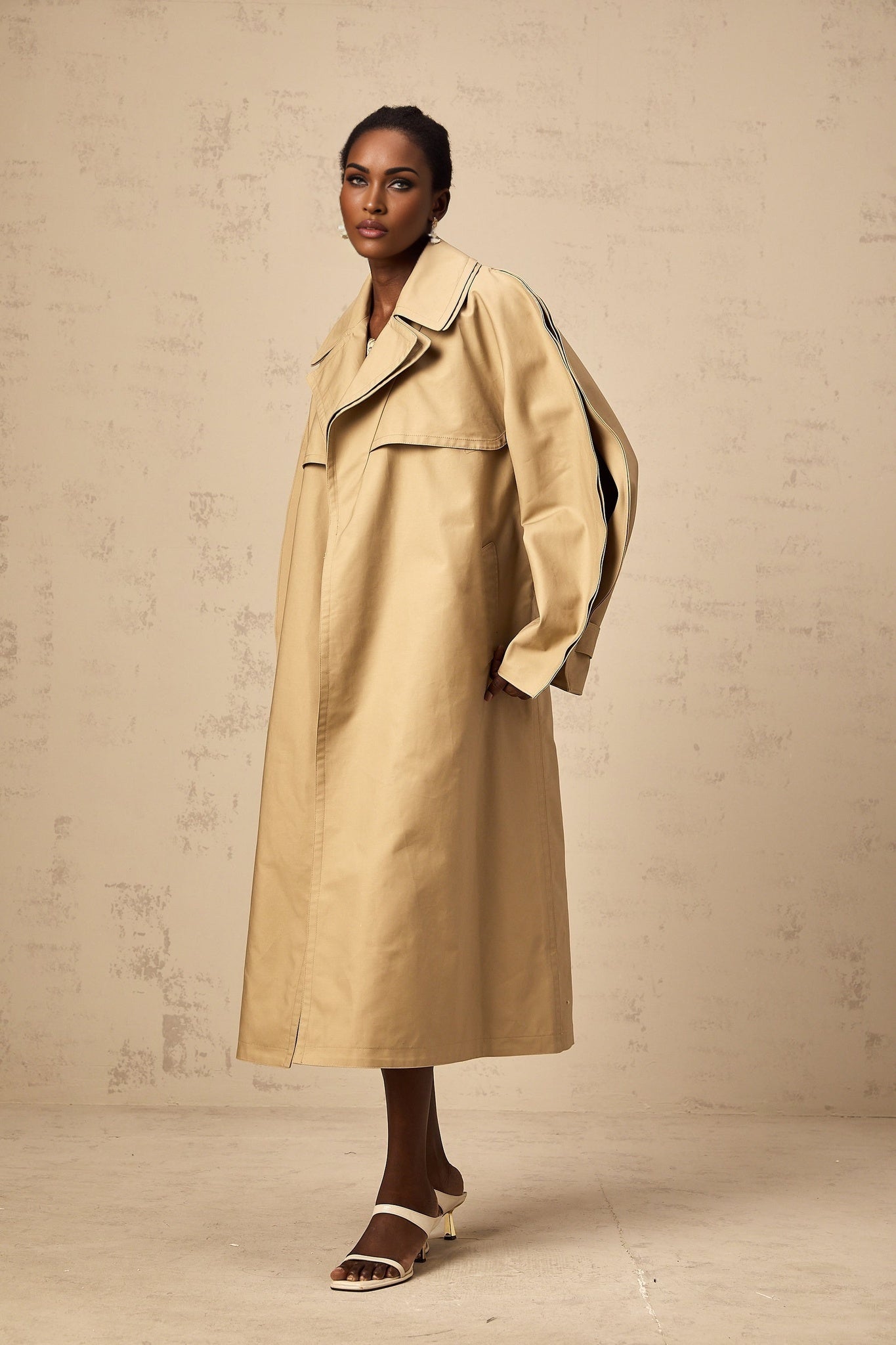 Fashionable khaki trench coat with curved sleeves worn by model standing against plain background