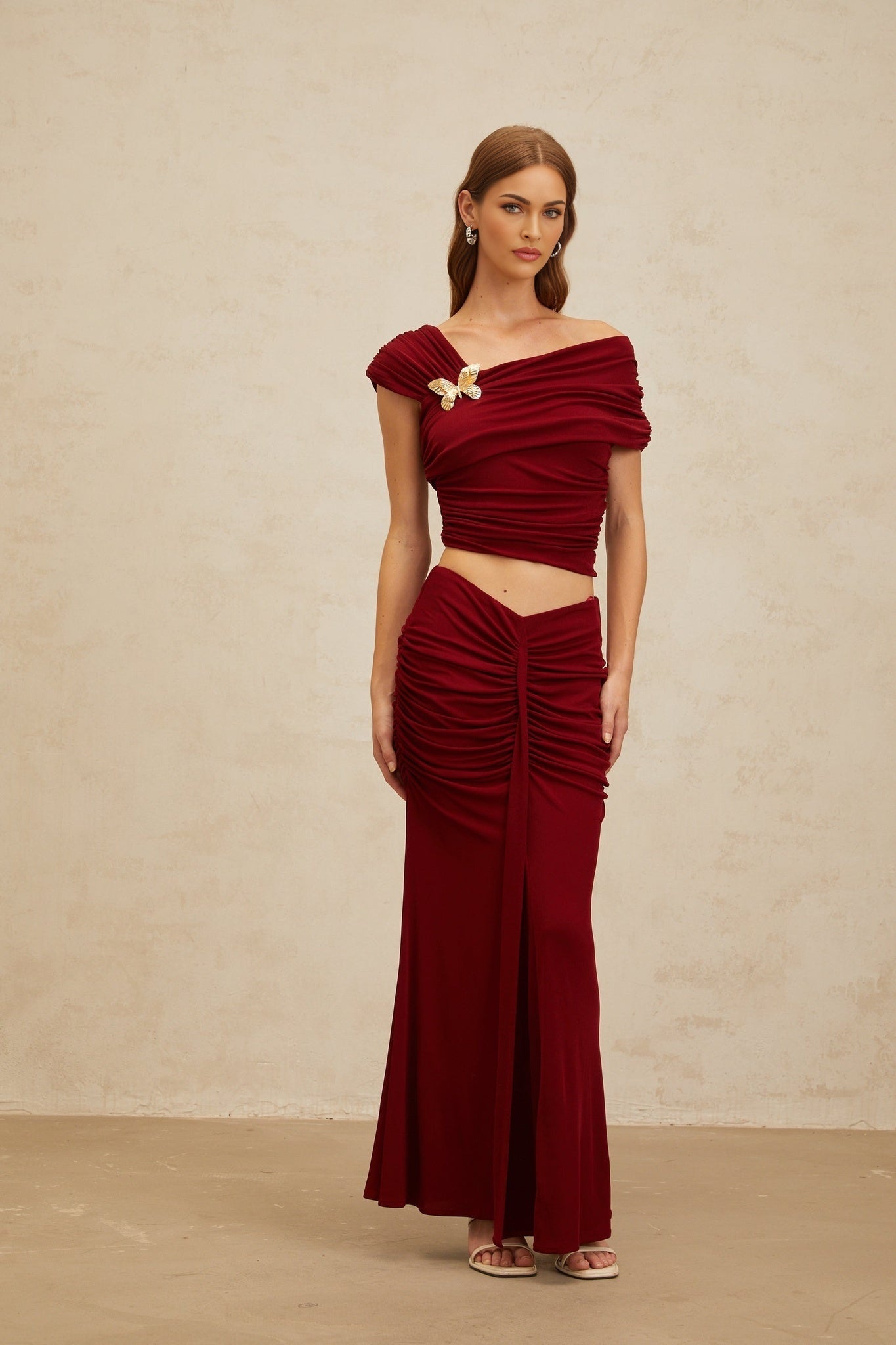 Red asymmetrical cropped top with butterfly brooch and cap sleeves, paired with red mermaid hem maxi skirt with front slit and ruched detailing