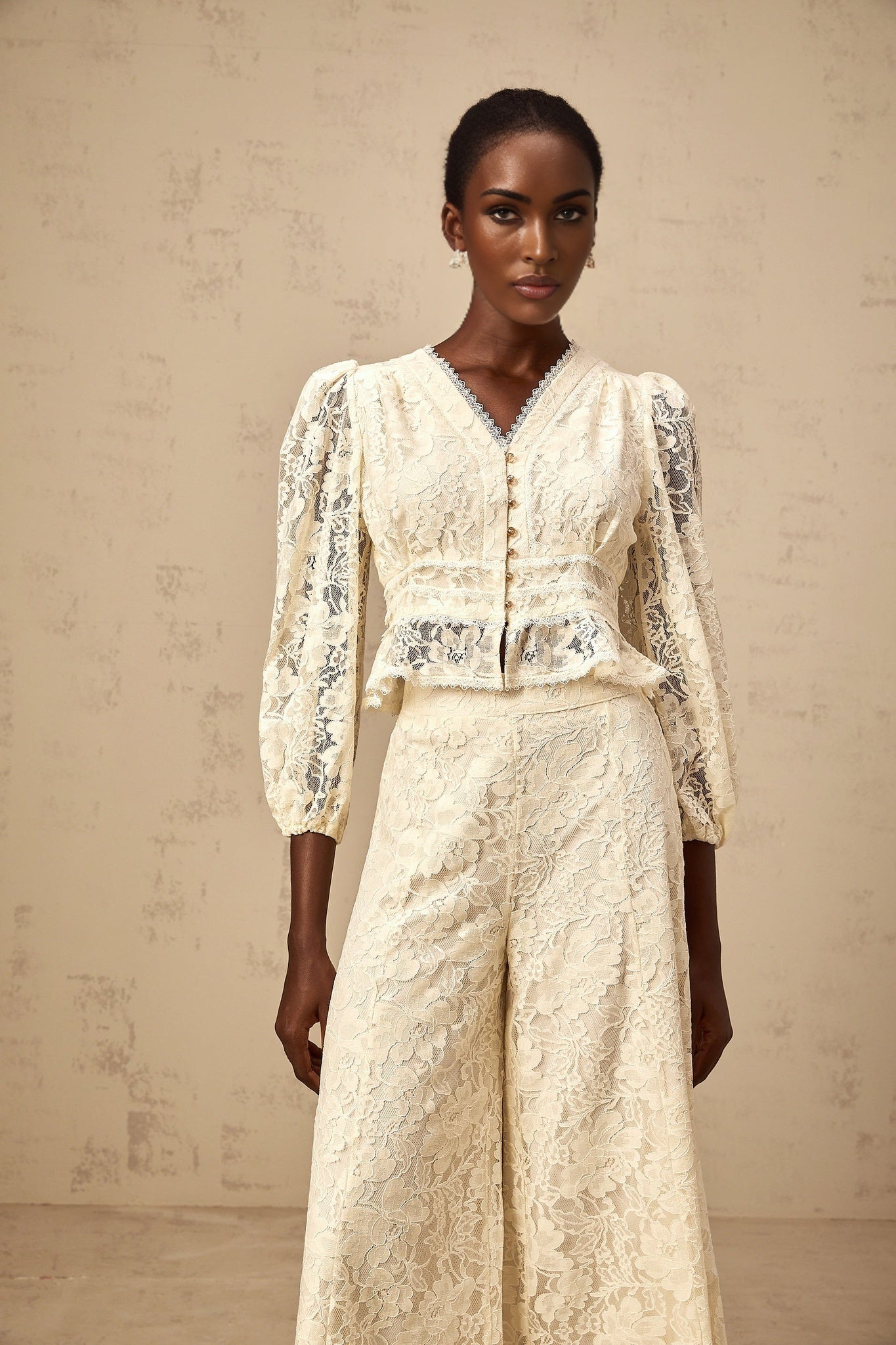 Woman wearing a beige floral lace blouse with matching trousers in a neutral color standing against a plain background