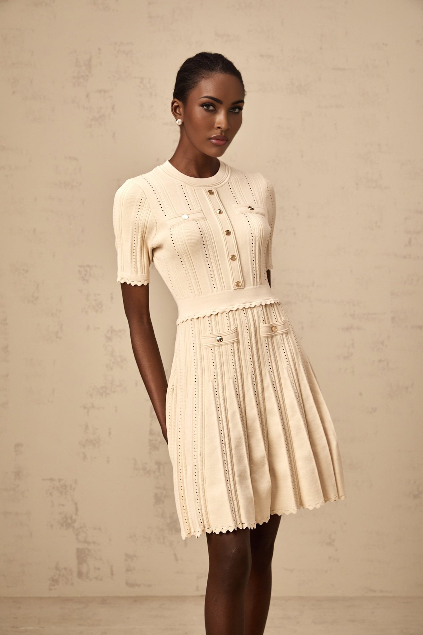 Woman wearing a beige mini dress with pleated skirt and delicate pointelle trim detail standing in a neutral setting