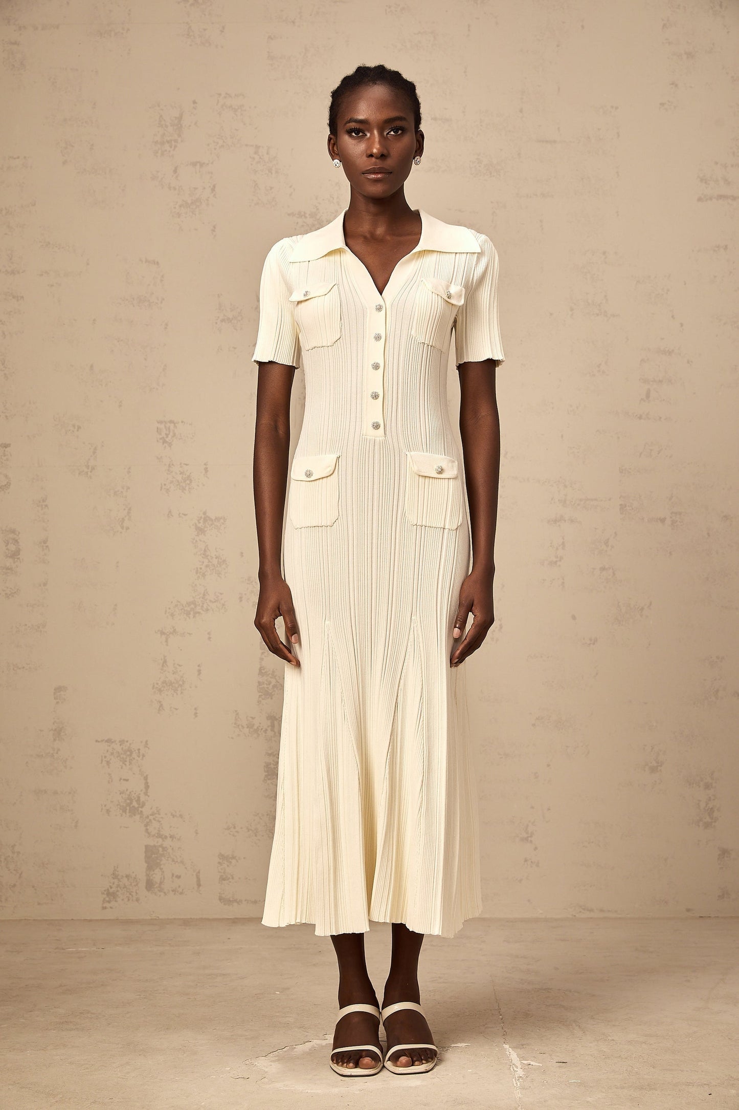 Woman wearing a beige ribbedknit midi dress with long sleeves and a formfitting silhouette standing against a plain background