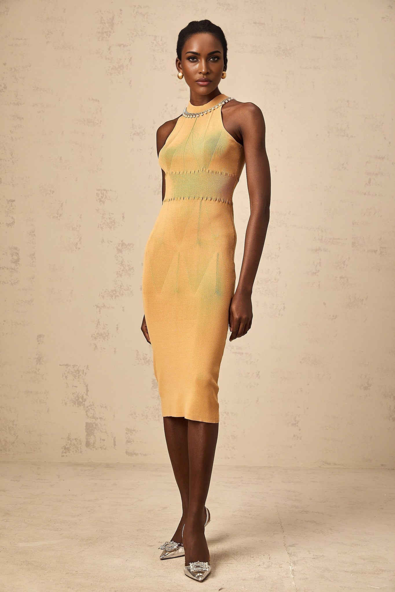 Woman wearing a cream colored knit midi dress with faux pearl embellishments on the neckline and sleeves standing in a neutral background