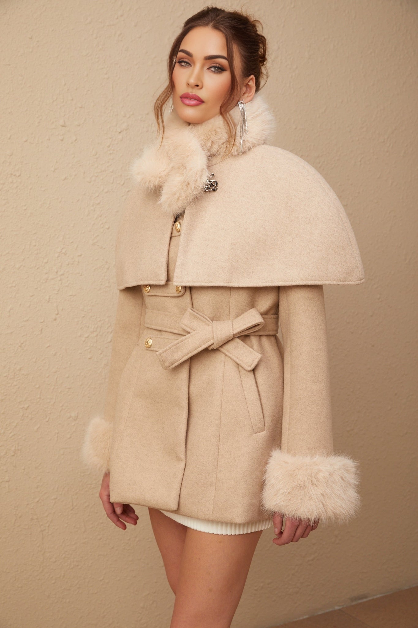 Woman wearing a long black faux fur coat with a detachable cape collar standing against a plain background
