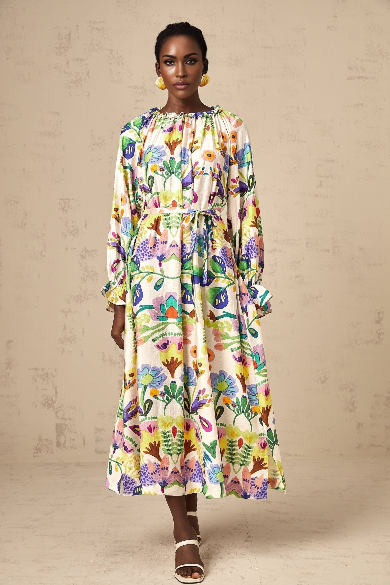 Woman wearing a long flowy midi dress with a vibrant paisley print pattern on light colored fabric standing in a casual pose