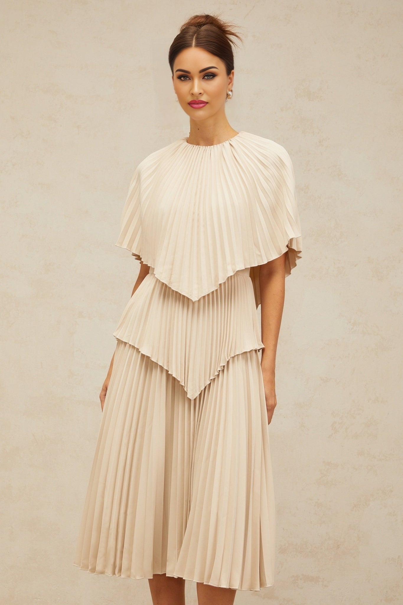 Woman wearing a neutral colored cape style pleated midi dress with flowing fabric and elegant drape