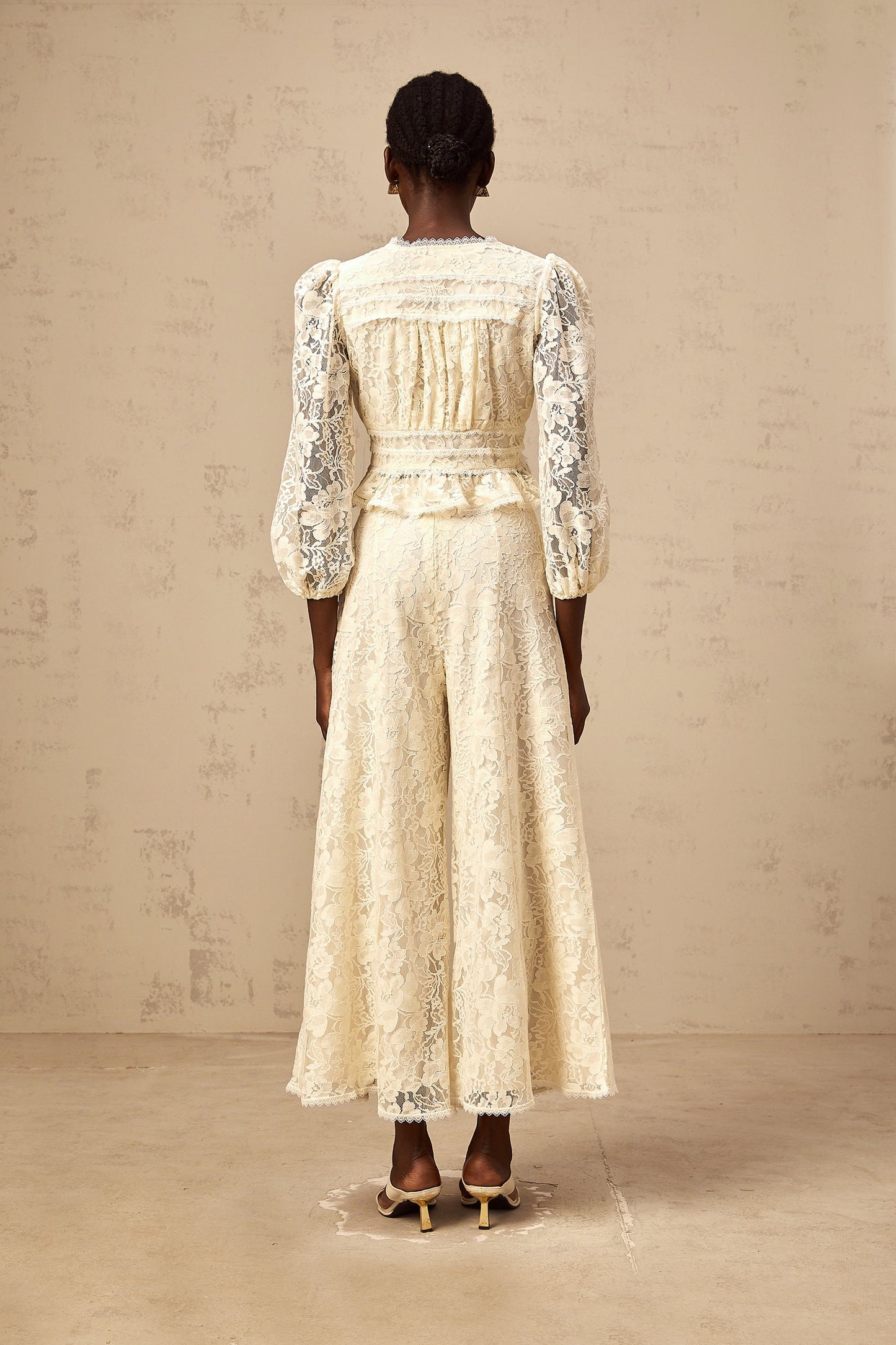 Woman wearing a neutral colored floral lace blouse and matching trousers set named Jacinthe standing against plain background