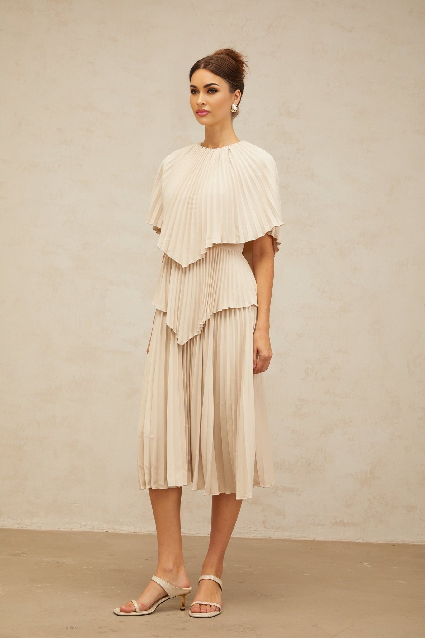 Woman wearing a neutral tone cape style pleated midi dress with flowy fabric and elegant draping