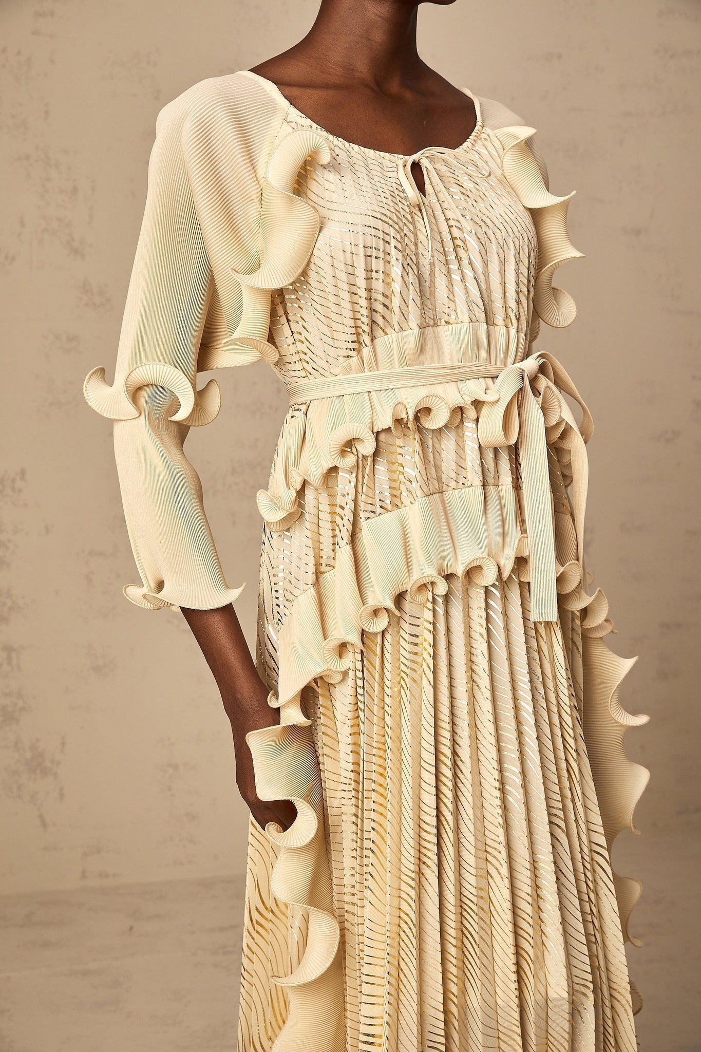 Woman wearing a neutralcolored midi dress with ruffled details and gold accents standing in a relaxed pose