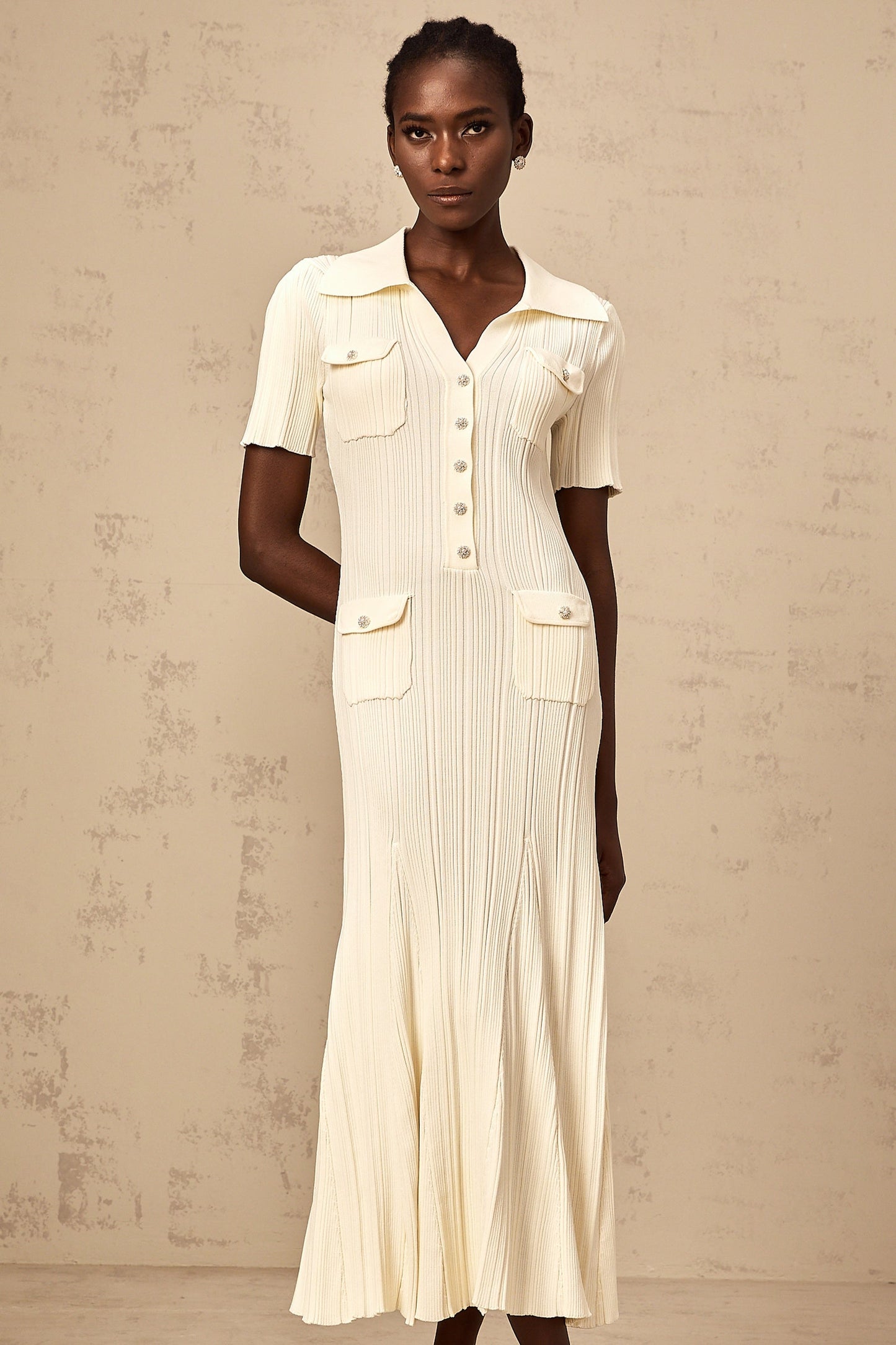 Woman wearing a neutralcolored ribbedknit midi dress with long sleeves and a formfitting silhouette standing against a plain background