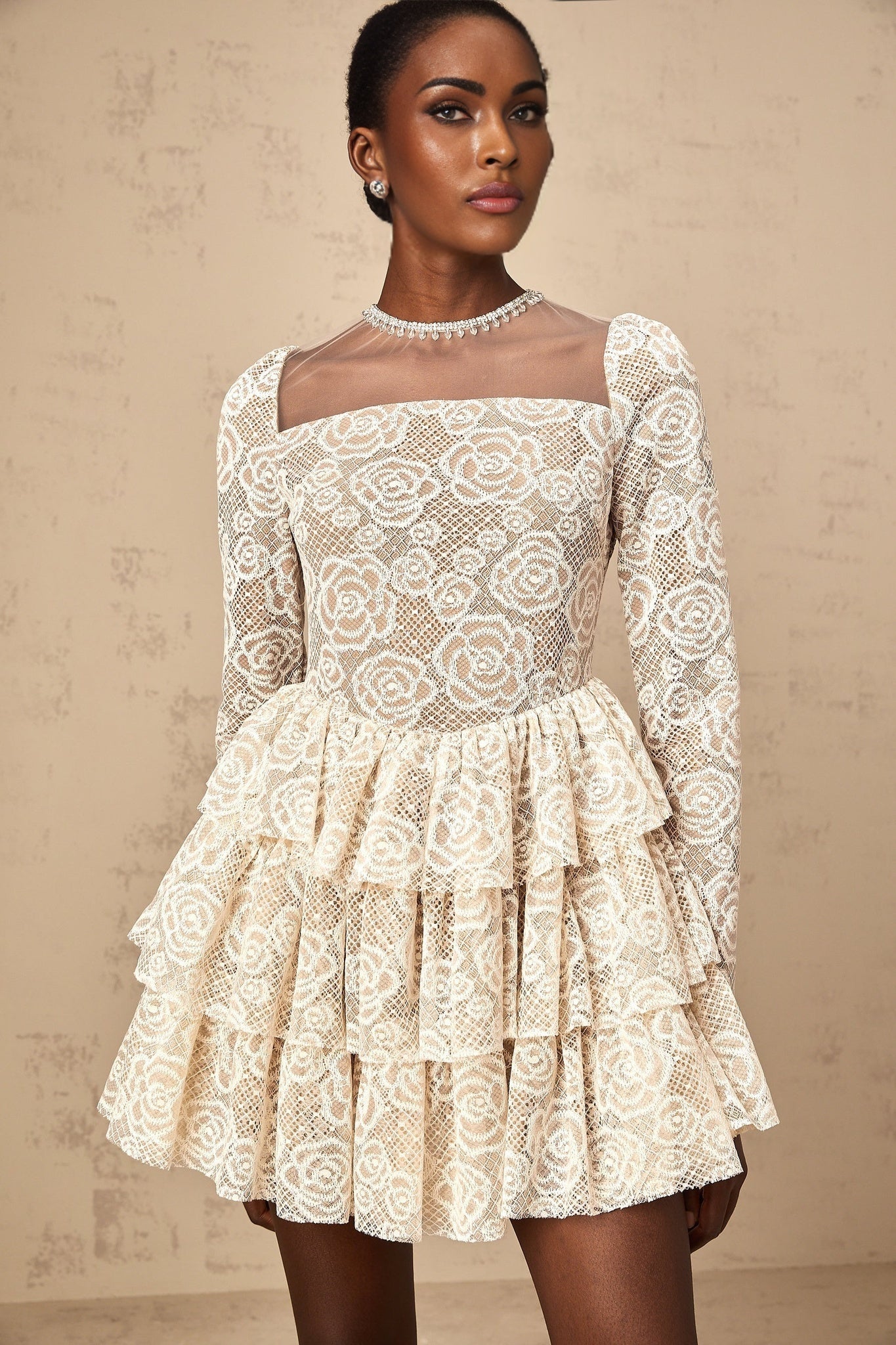 Woman wearing a short beige dress with floral embroidery details standing against a plain background