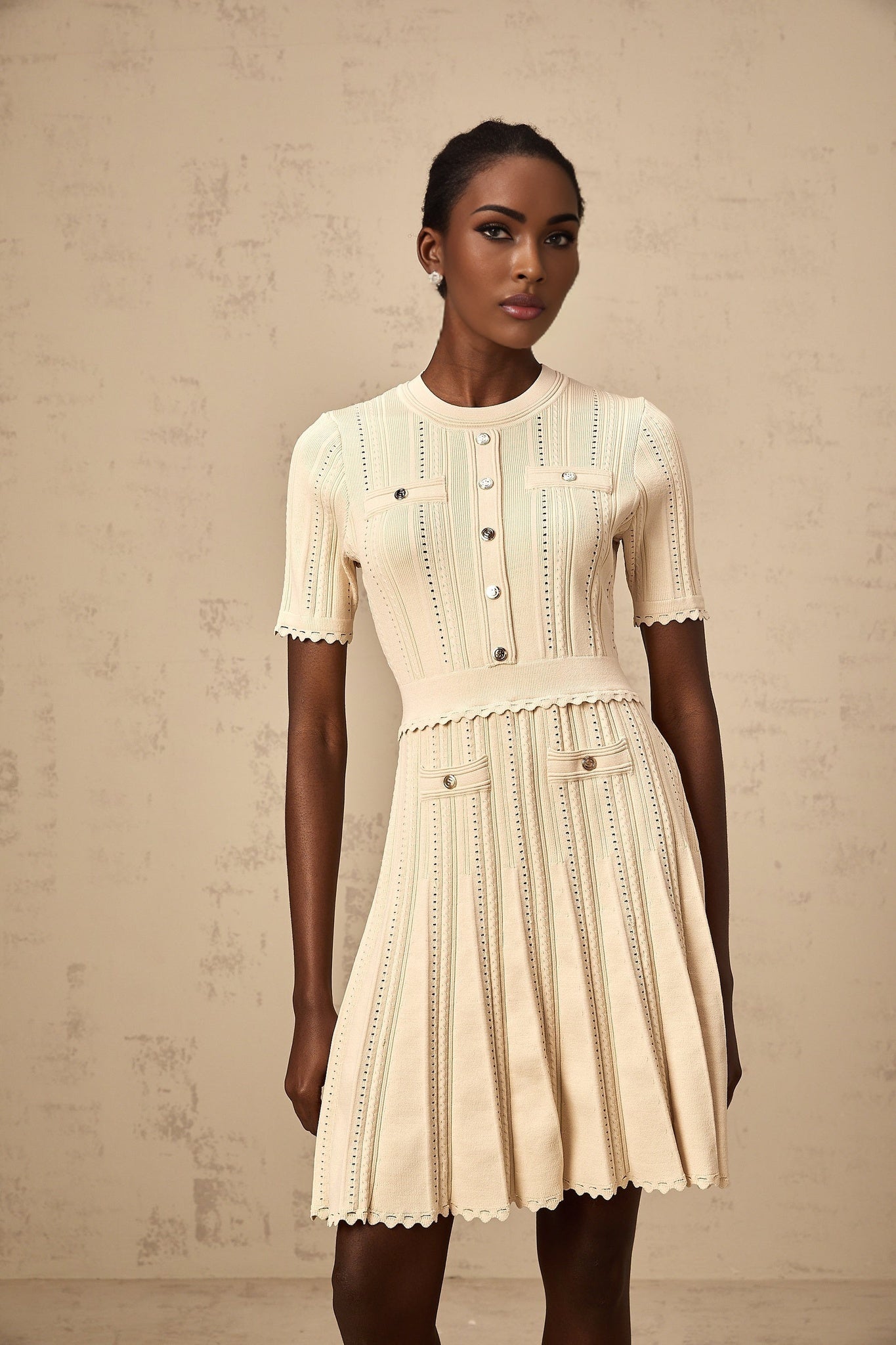Woman wearing a short beige knitted dress with pleated skirt and delicate pointelle trim details standing in a neutral setting