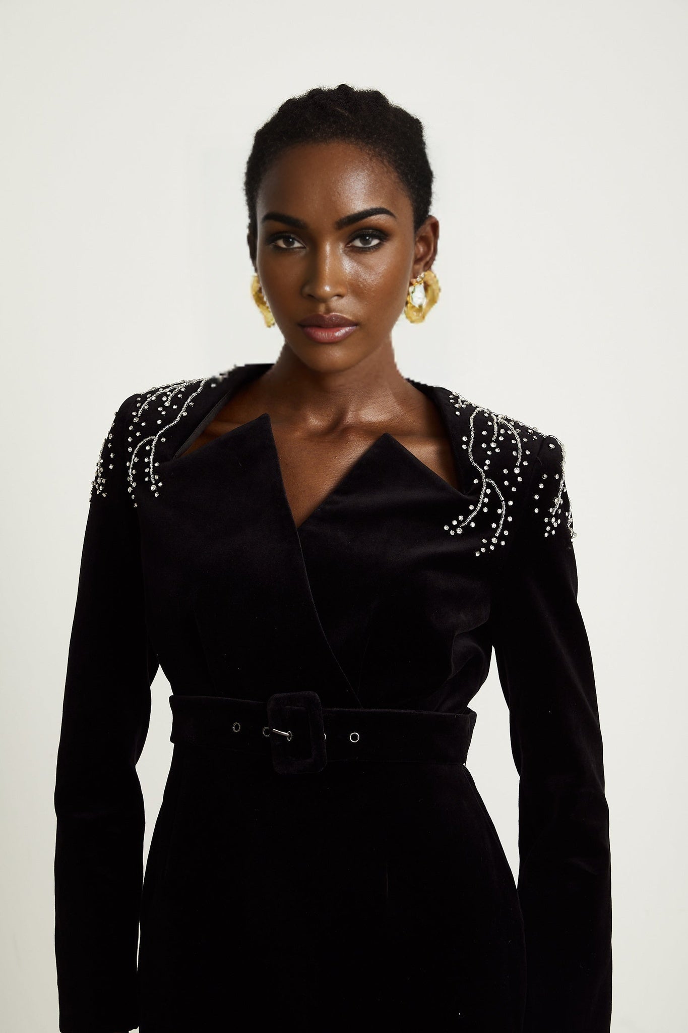 Woman wearing a short black velvet dress with crystal embellishments and thin straps standing in front of plain background
