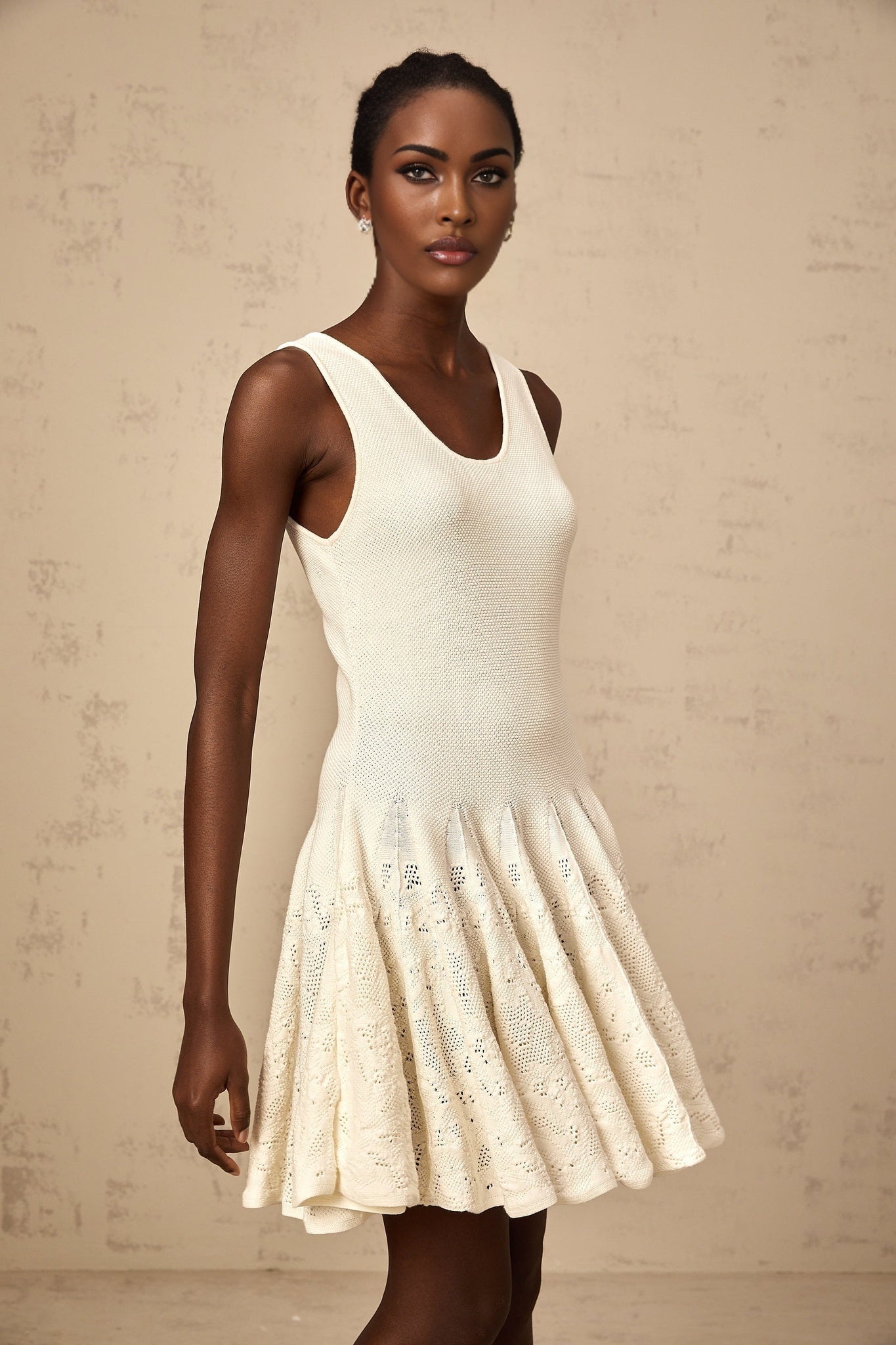 Woman wearing a short white textured knit dress with intricate jacquard pattern standing against plain background