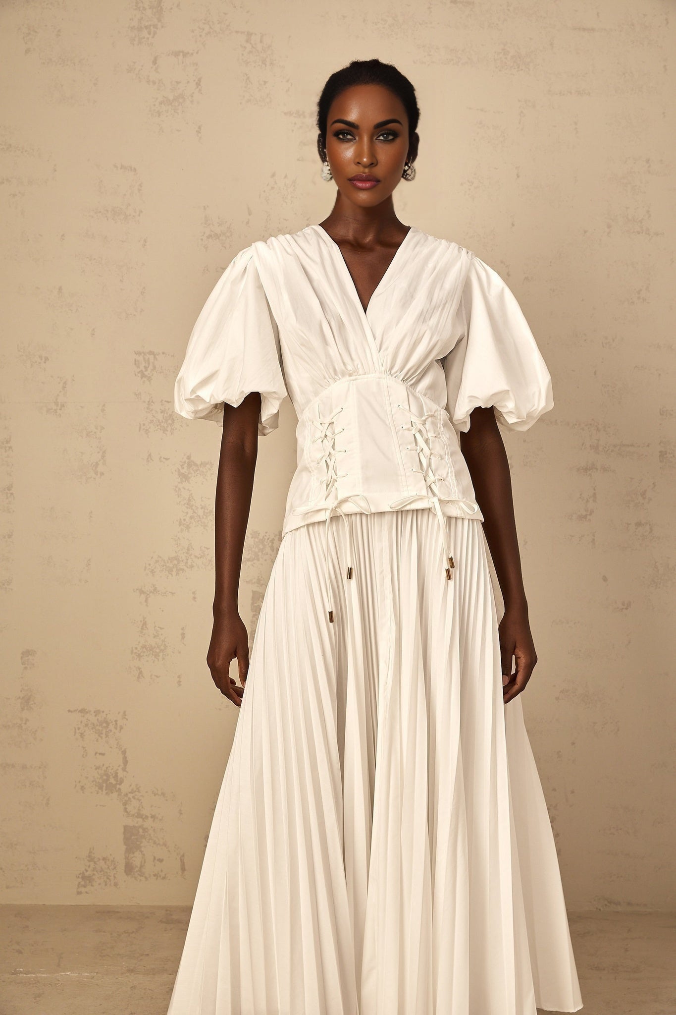 Woman wearing a white laceup pleated maxi dress with long sleeves and a flowy skirt standing against a plain background