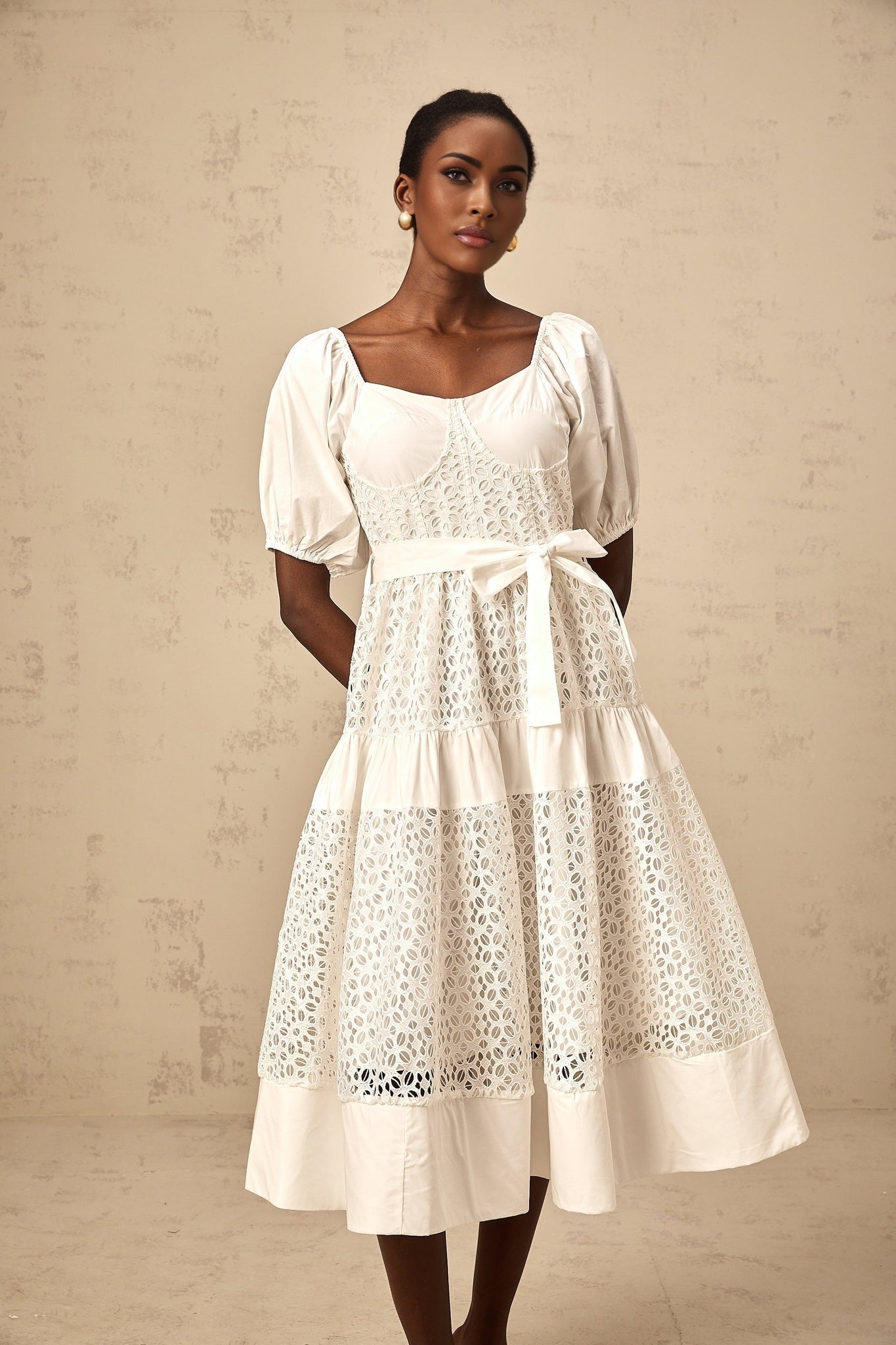 Woman wearing a white tiered midi dress made of corded lace with long sleeves and a high neckline standing against a plain background