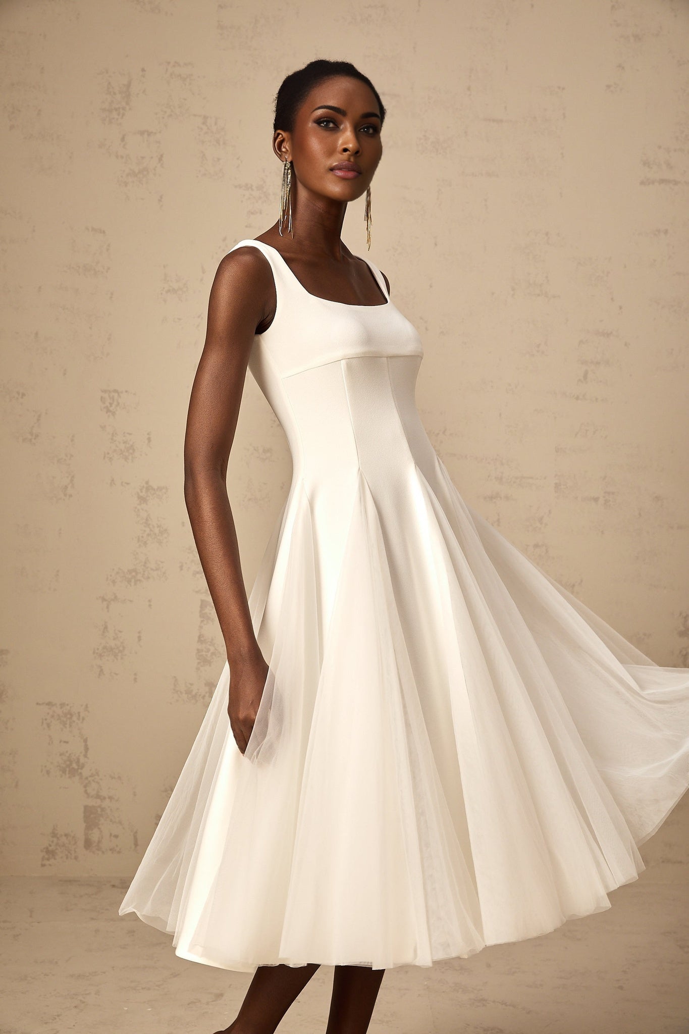 Woman wearing a white tulle midi dress with godet inserts and flared skirt standing in a neutral background