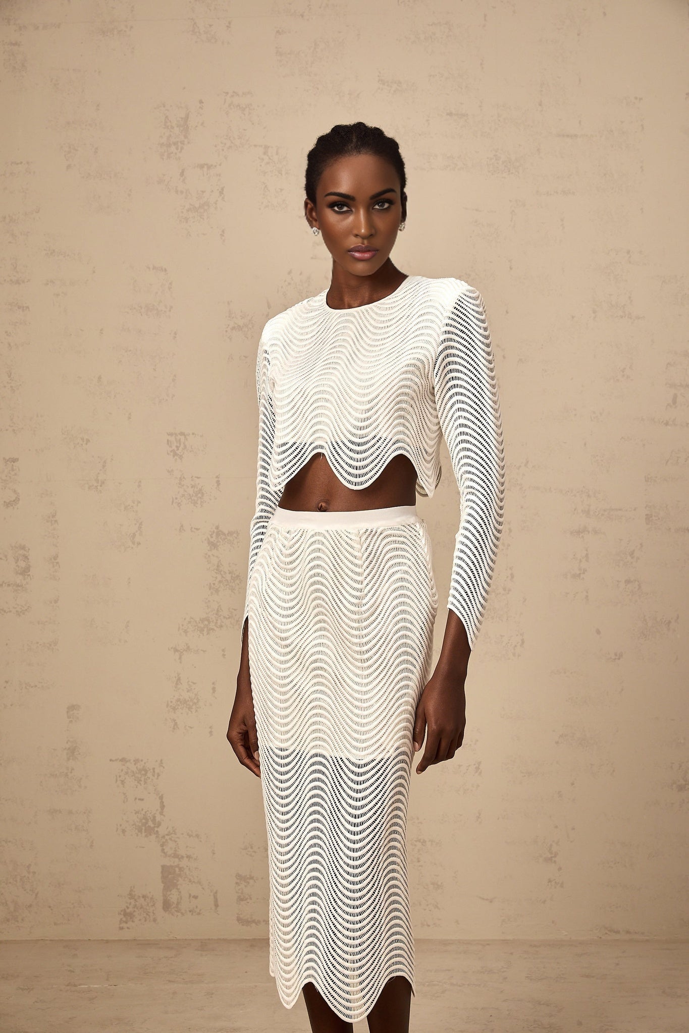Woman wearing a white twopiece outfit consisting of a wavy woven top and matching skirt standing against a plain background
