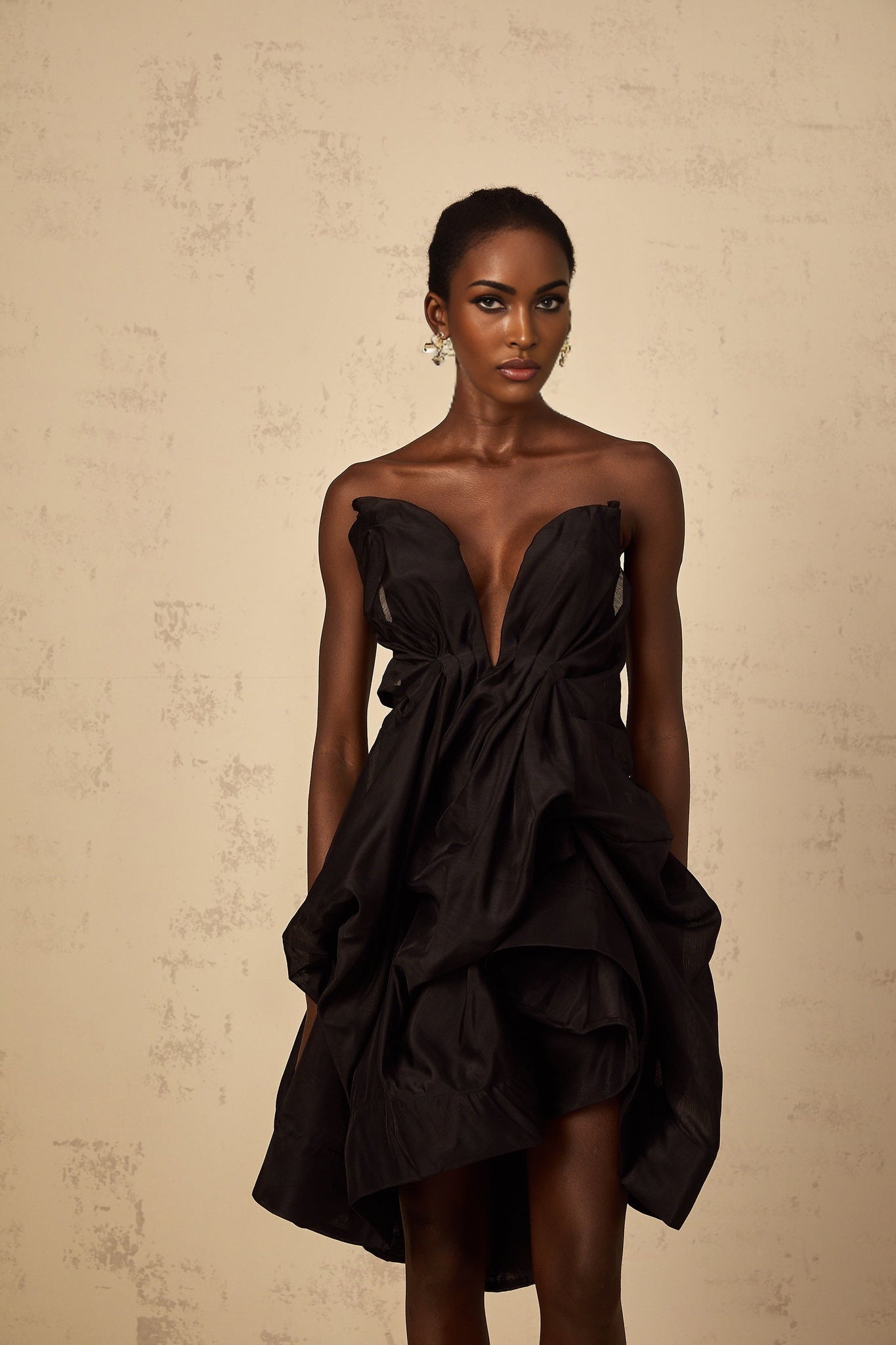 Woman wearing asymmetrical pleated cocktail mini dress in dark color standing against plain background