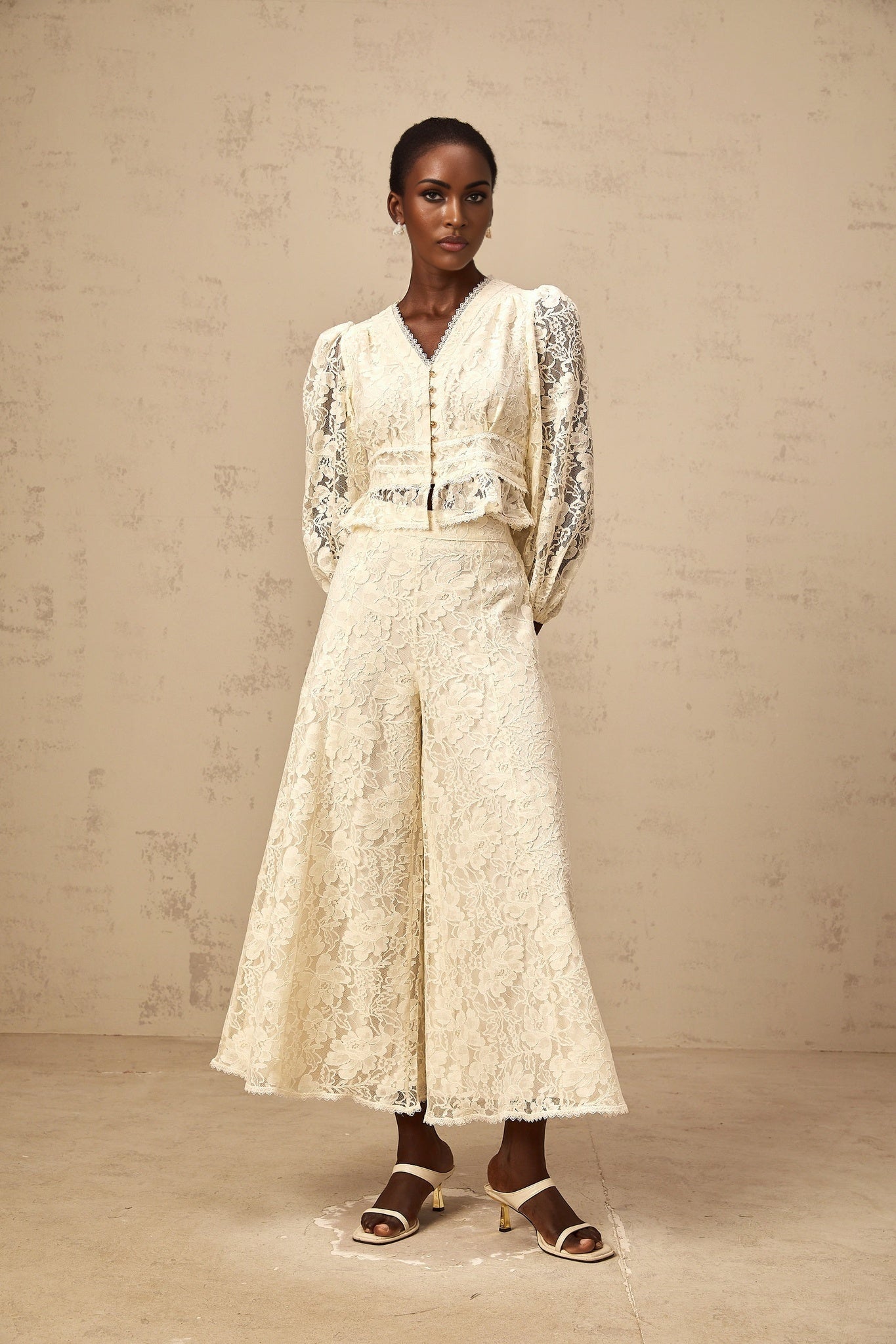 Woman wearing beige floral lace blouse and matching trousers set standing against plain background