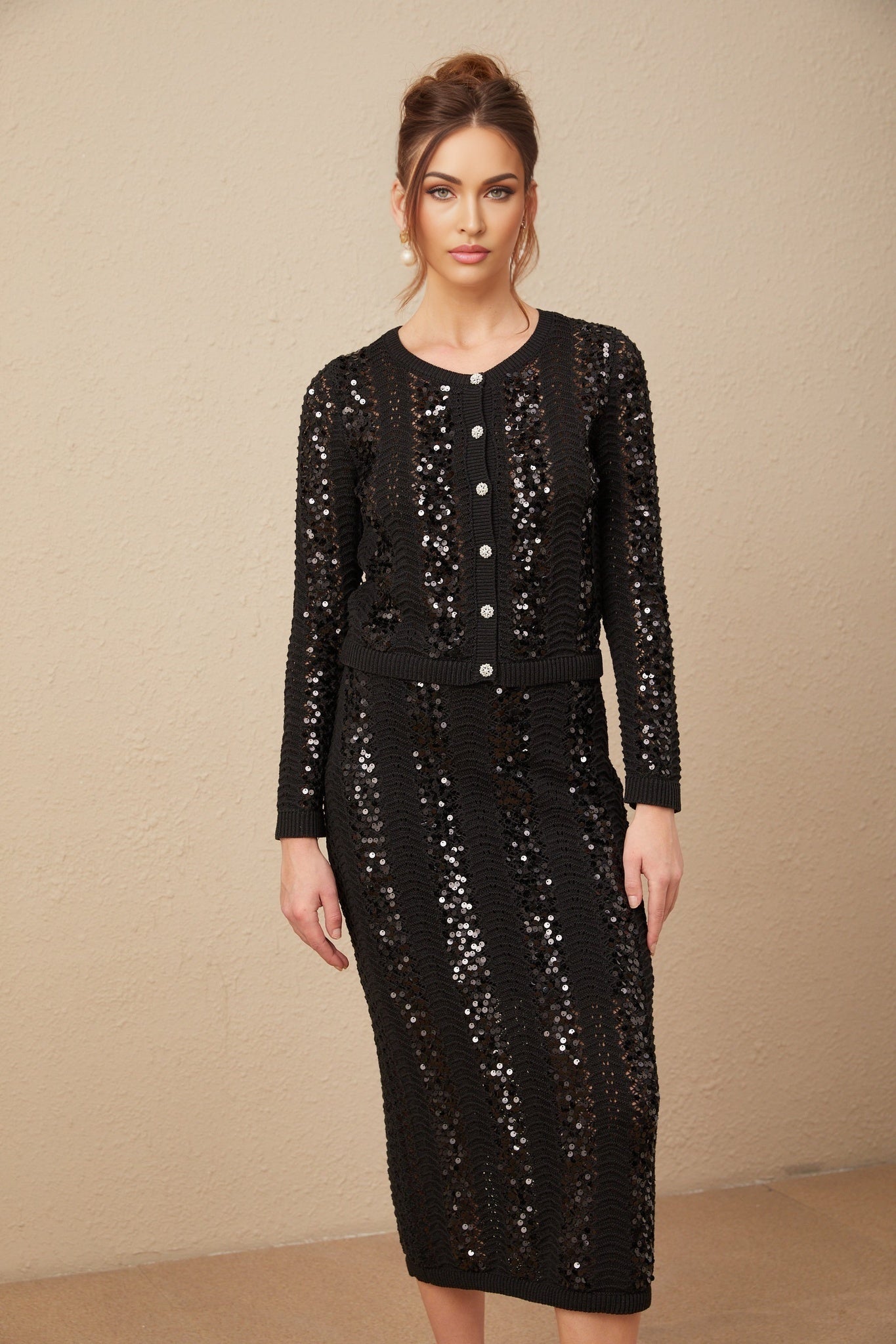 Woman wearing black sequined crochet knit cardigan and matching skirt set standing against plain background