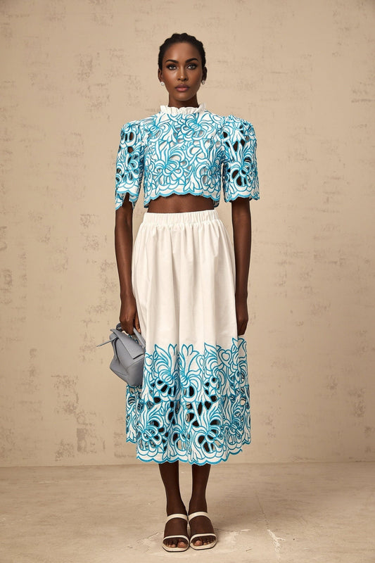 Woman wearing blue embroidered crop top and matching skirt with cutout details standing against light background