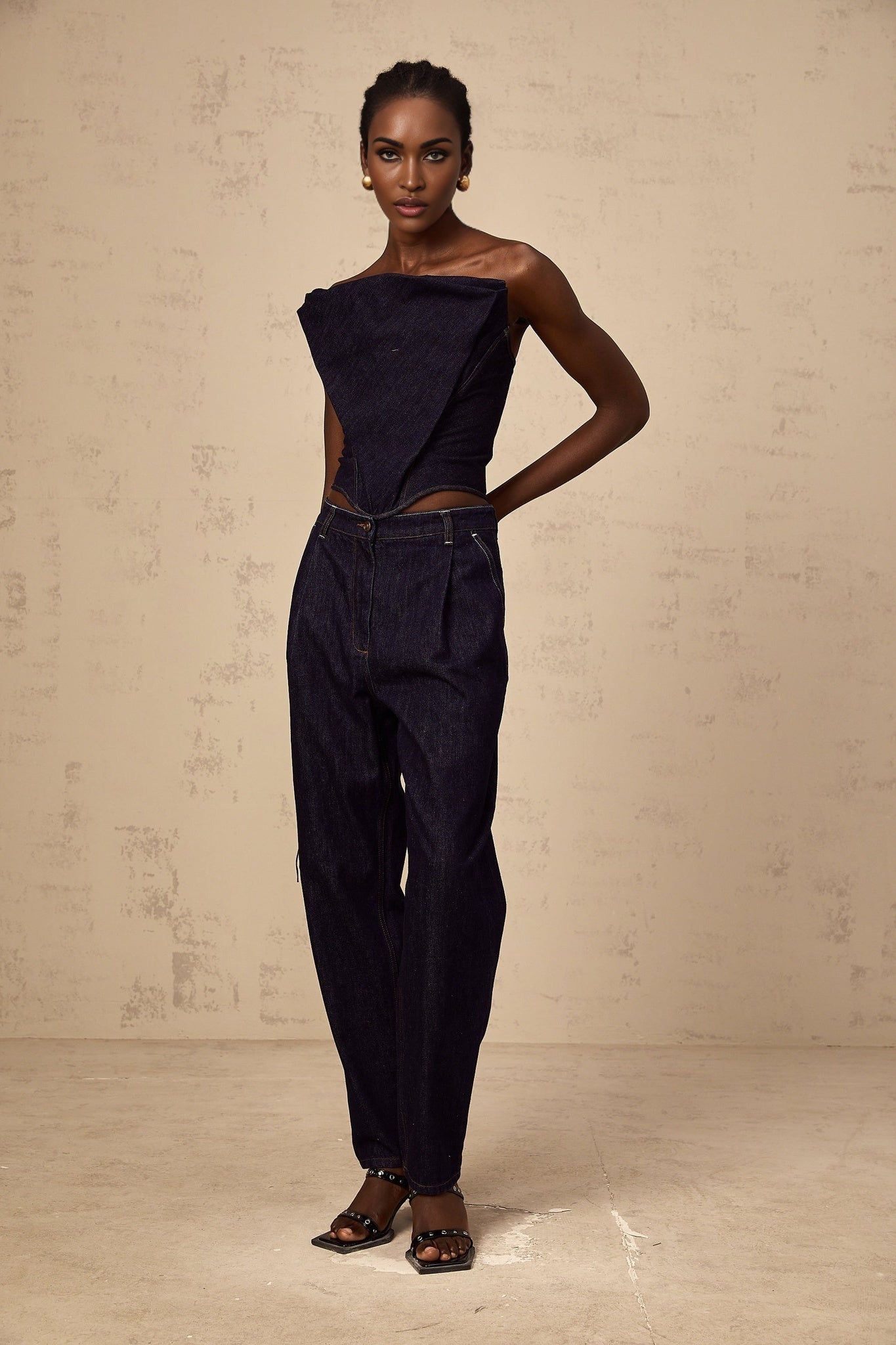 Woman wearing blue high rise jeans with pleated details on the front standing against a plain background