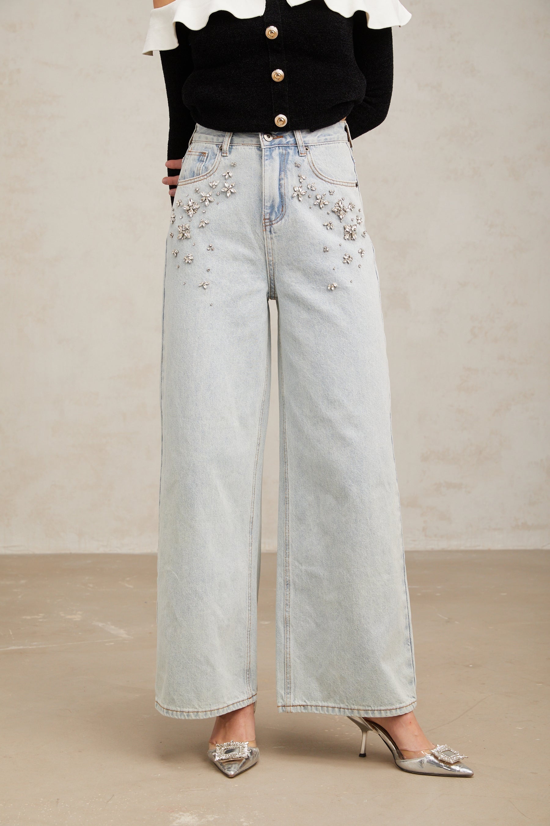 Woman wearing light blue jeans with decorative embellishments on the legs standing against a plain background