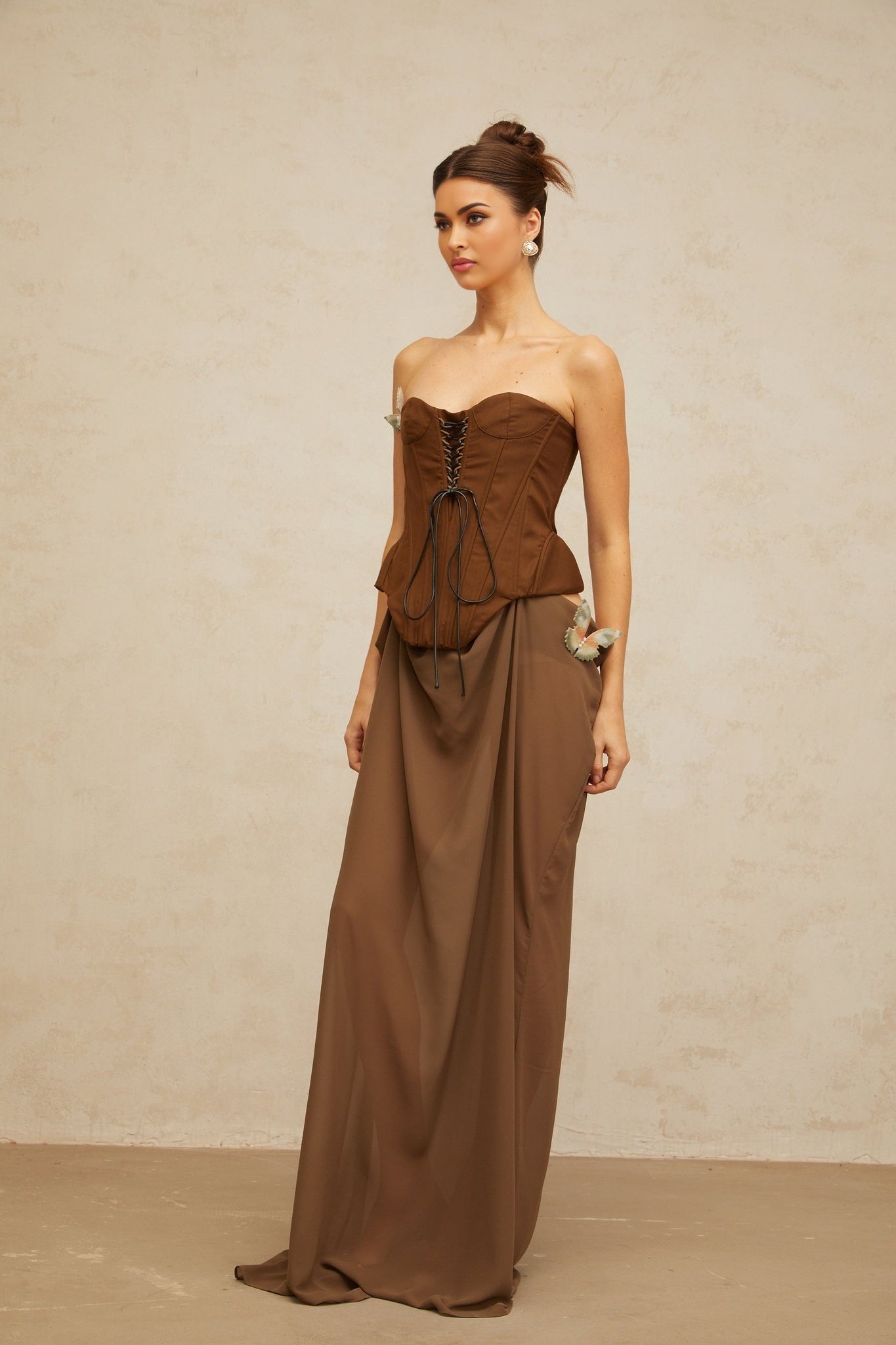 Woman wearing long brown corset maxi dress with butterfly pin detail and flowing skirt