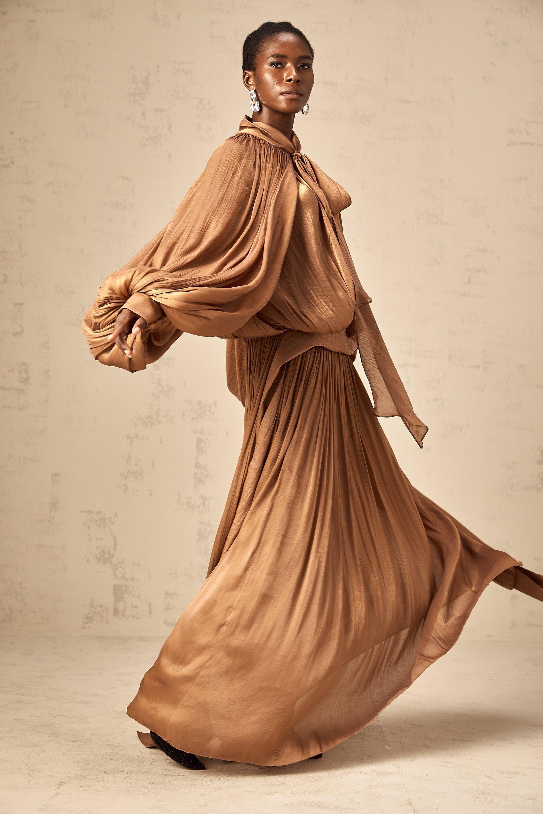 Woman wearing long brown pleated satin maxi dress with flowing skirt and fitted bodice
