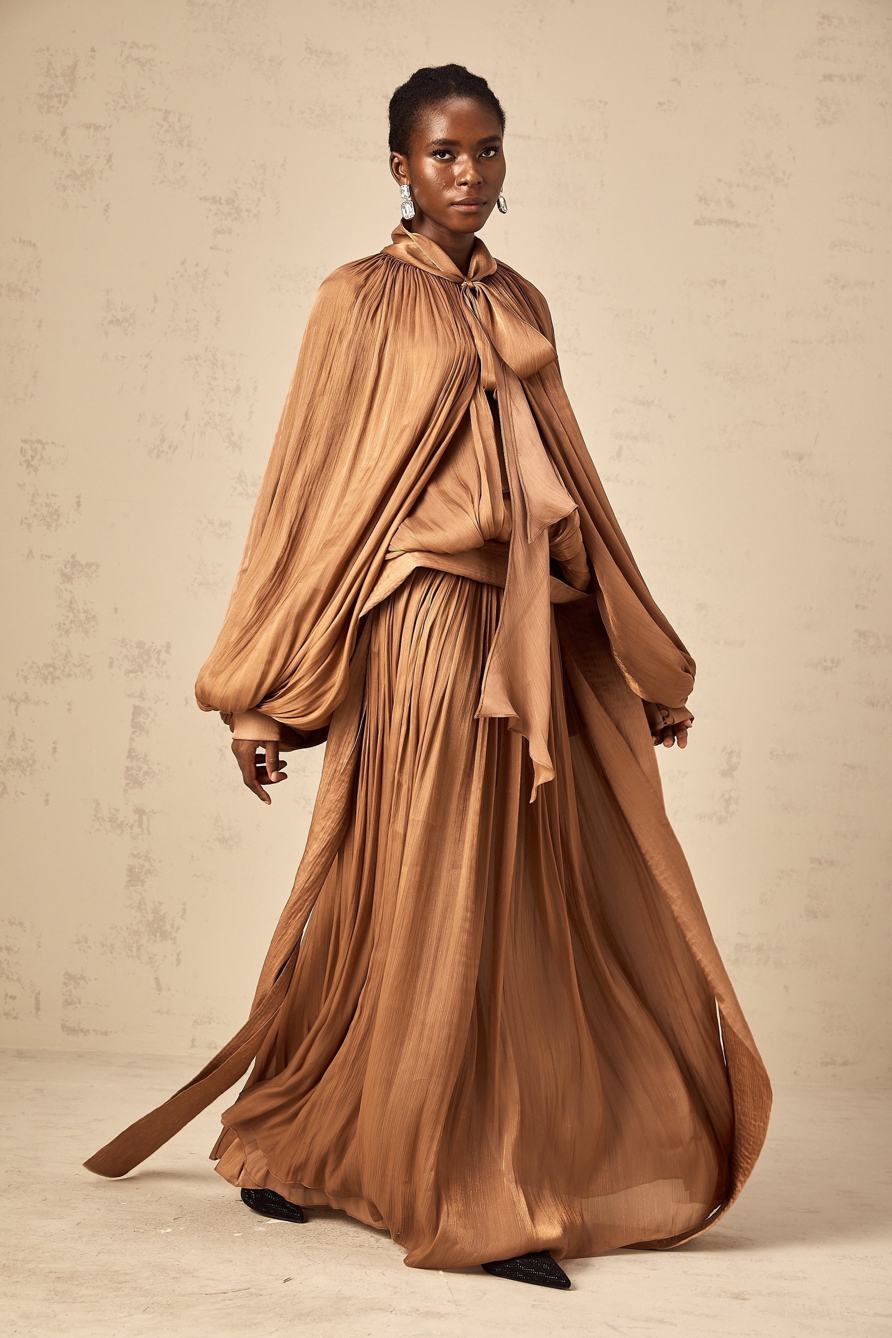 Woman wearing long brown satin maxi dress with pleated design flowing to the floor