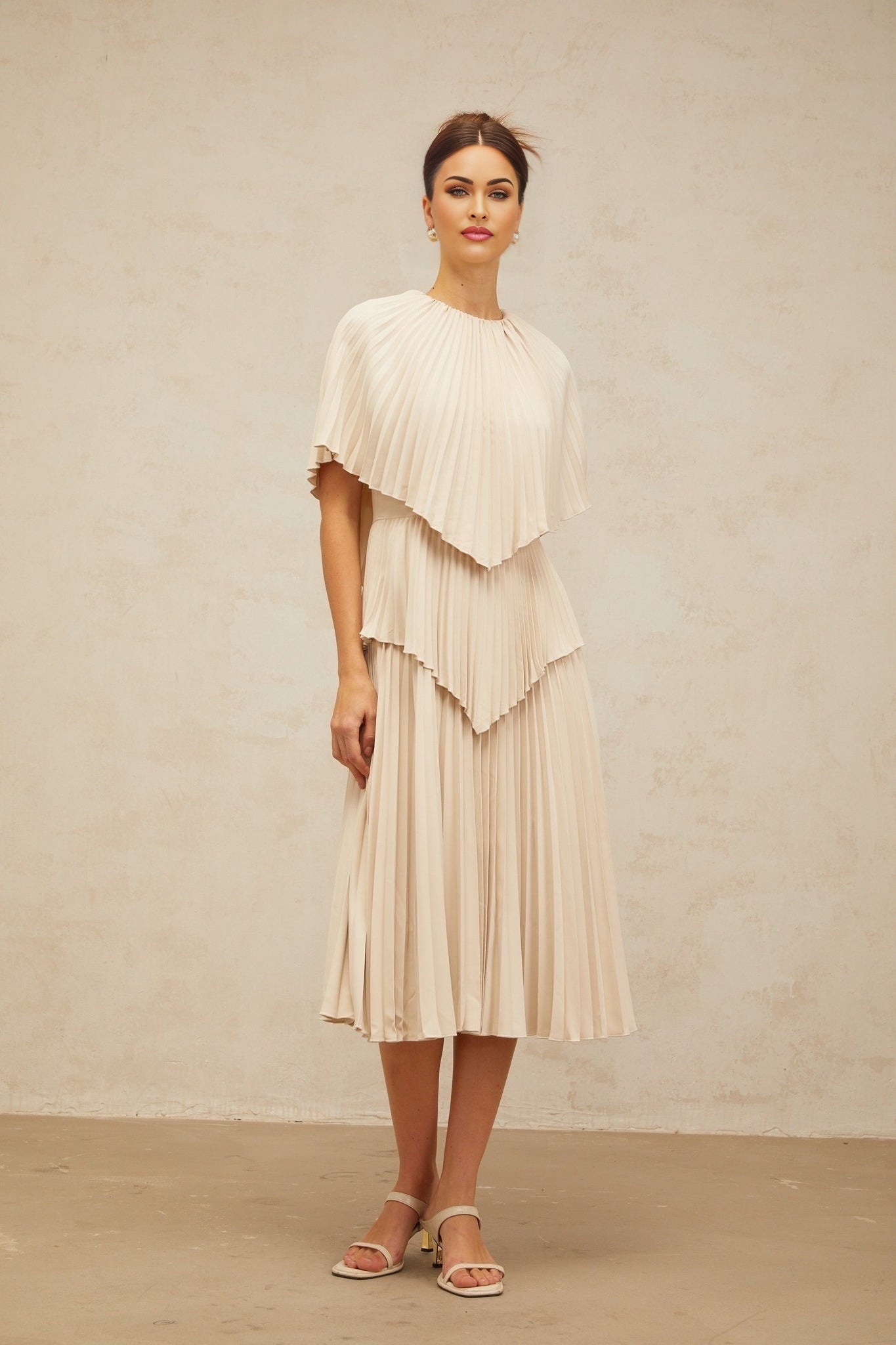 Woman wearing long flowing neutral colored pleated midi dress with cape detail