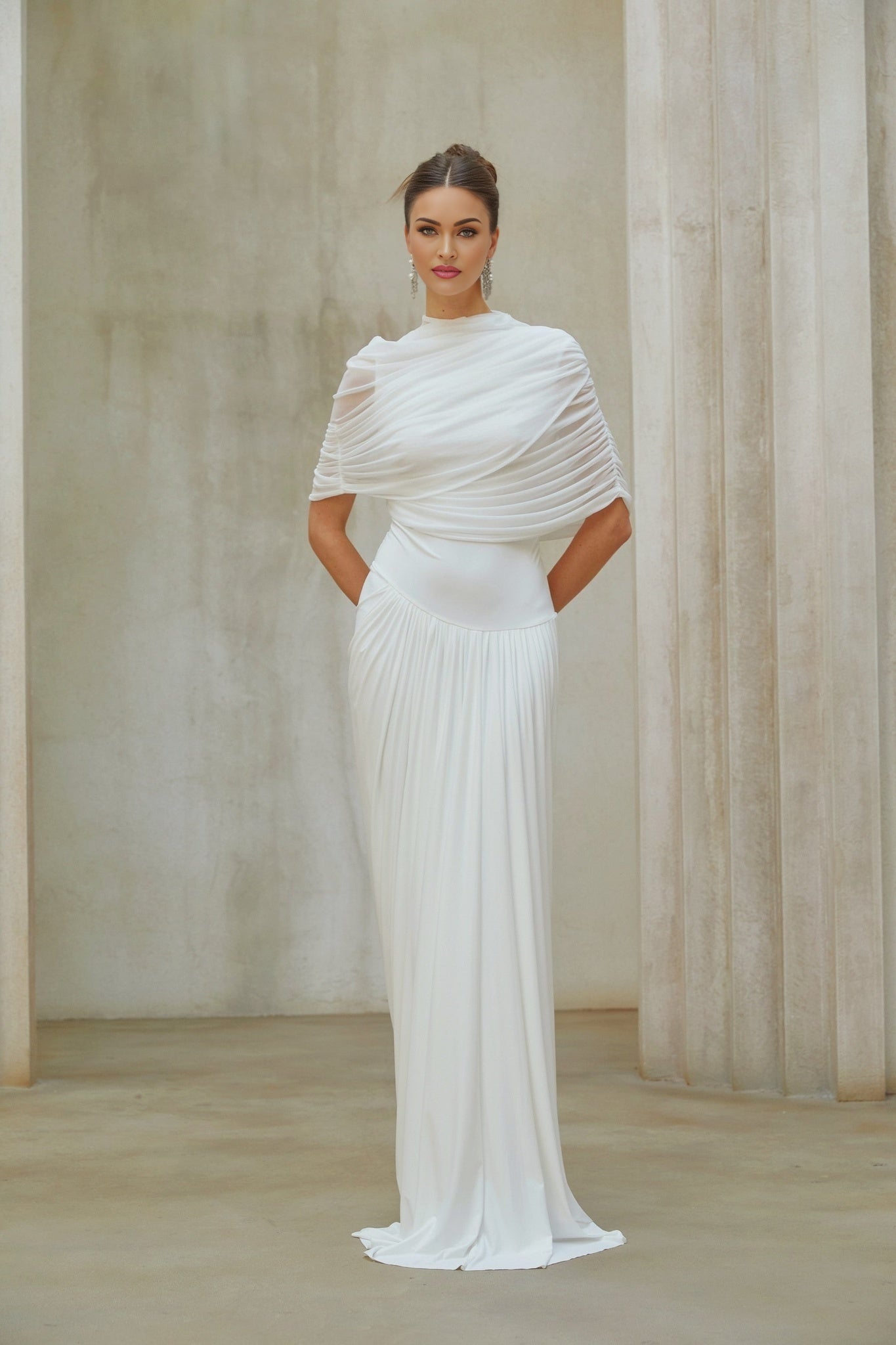 Woman wearing long white asymmetrical maxi dress with pleated details standing against plain background
