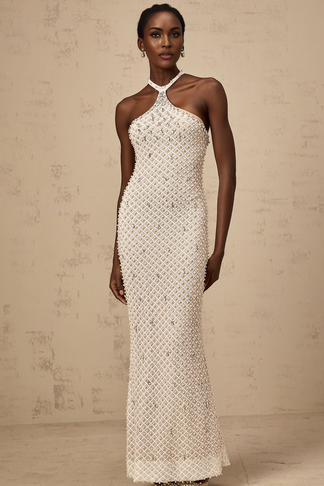 Woman wearing long white halterneck maxi dress with embellished details standing against plain background