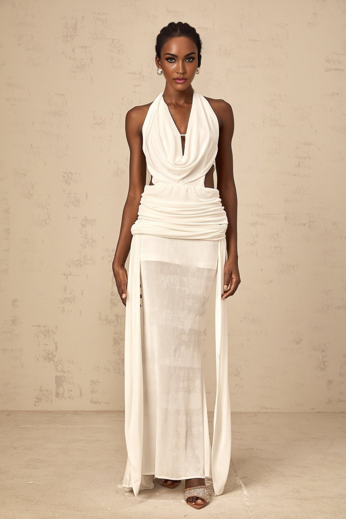 Woman wearing long white halterneck maxi dress with open back standing in front of plain background