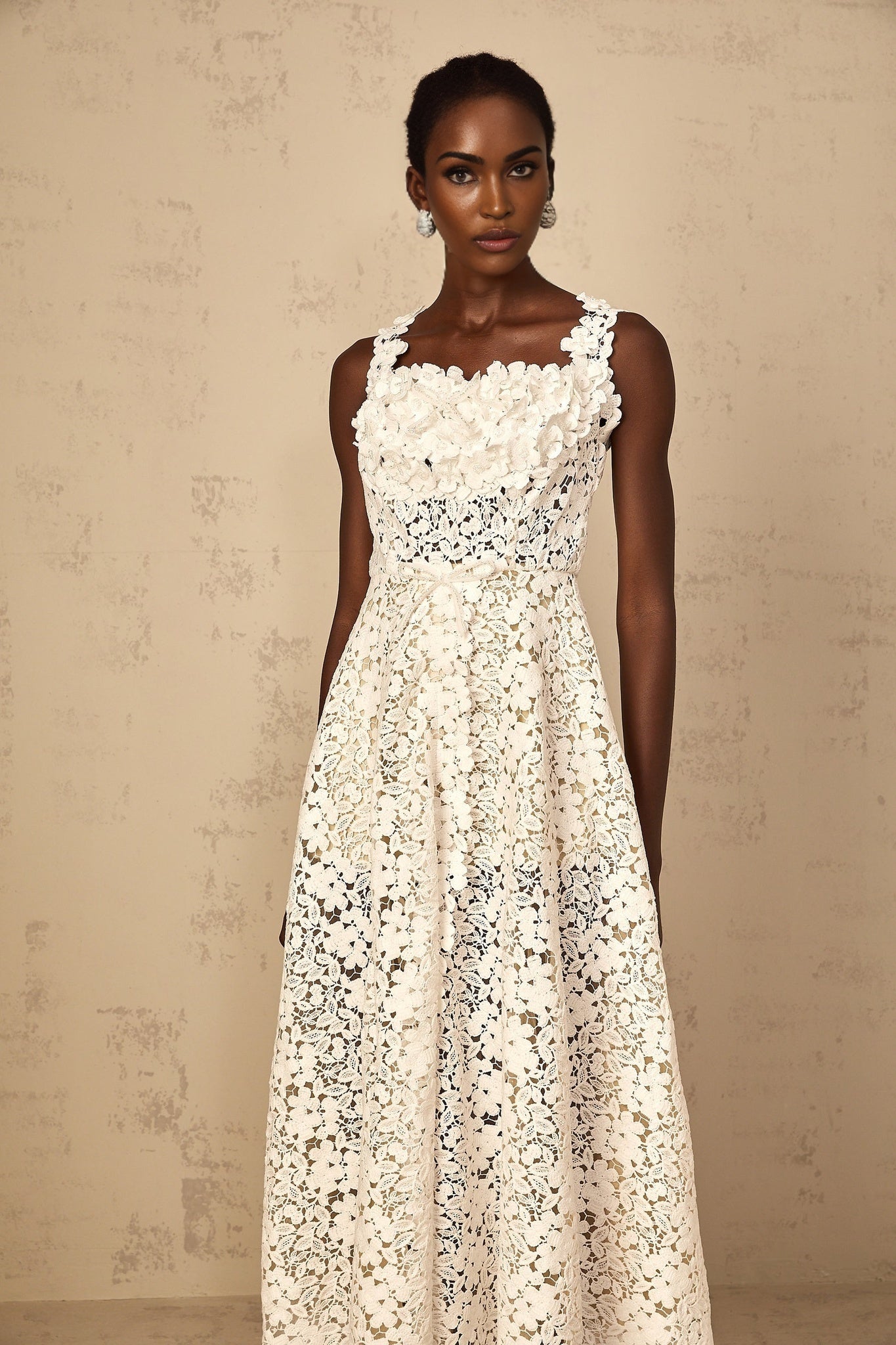 Woman wearing long white lace maxi dress with shirred details and corded lace pattern standing against plain background