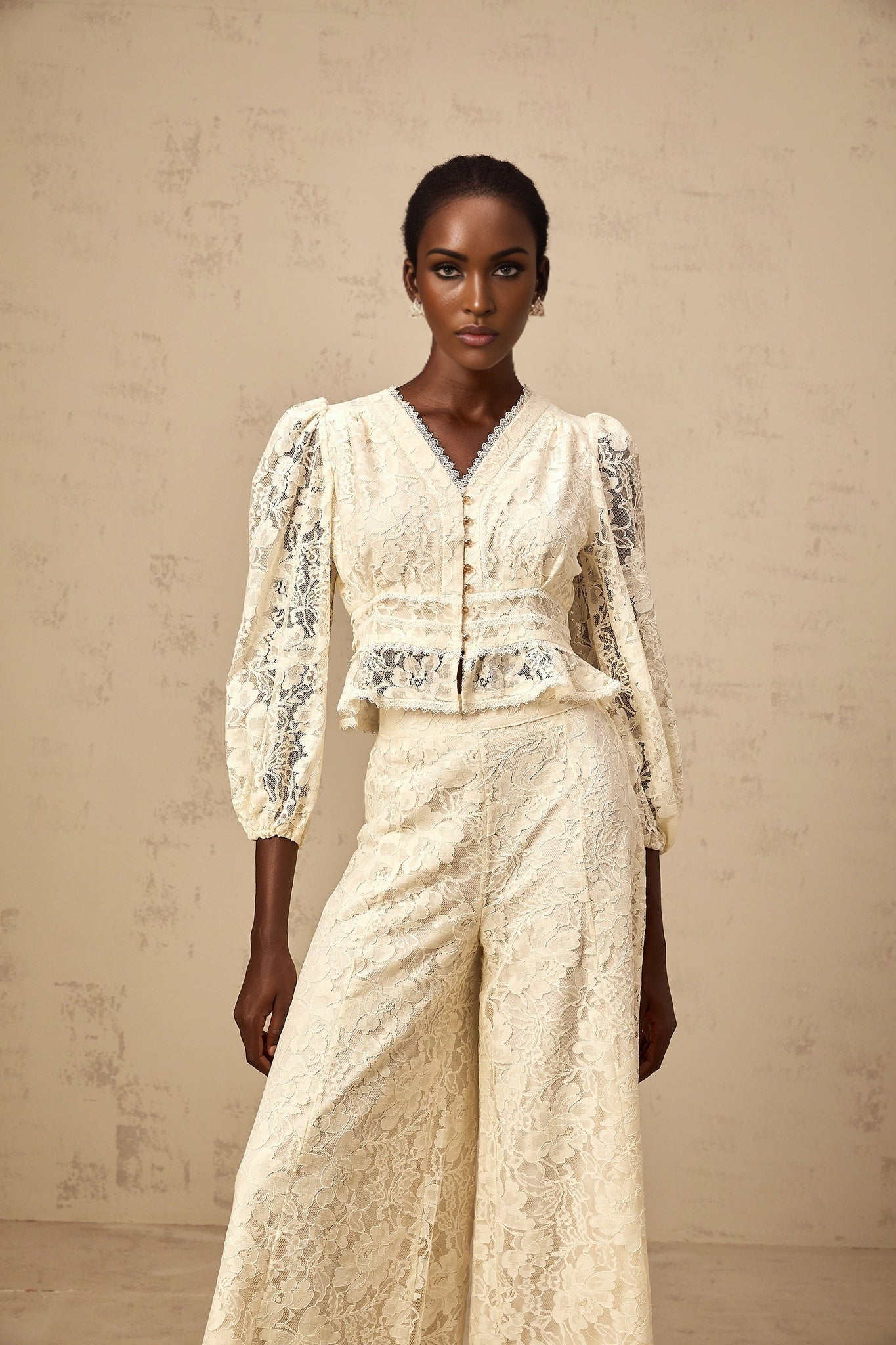 Woman wearing matching beige floral lace blouse and trousers set against plain background