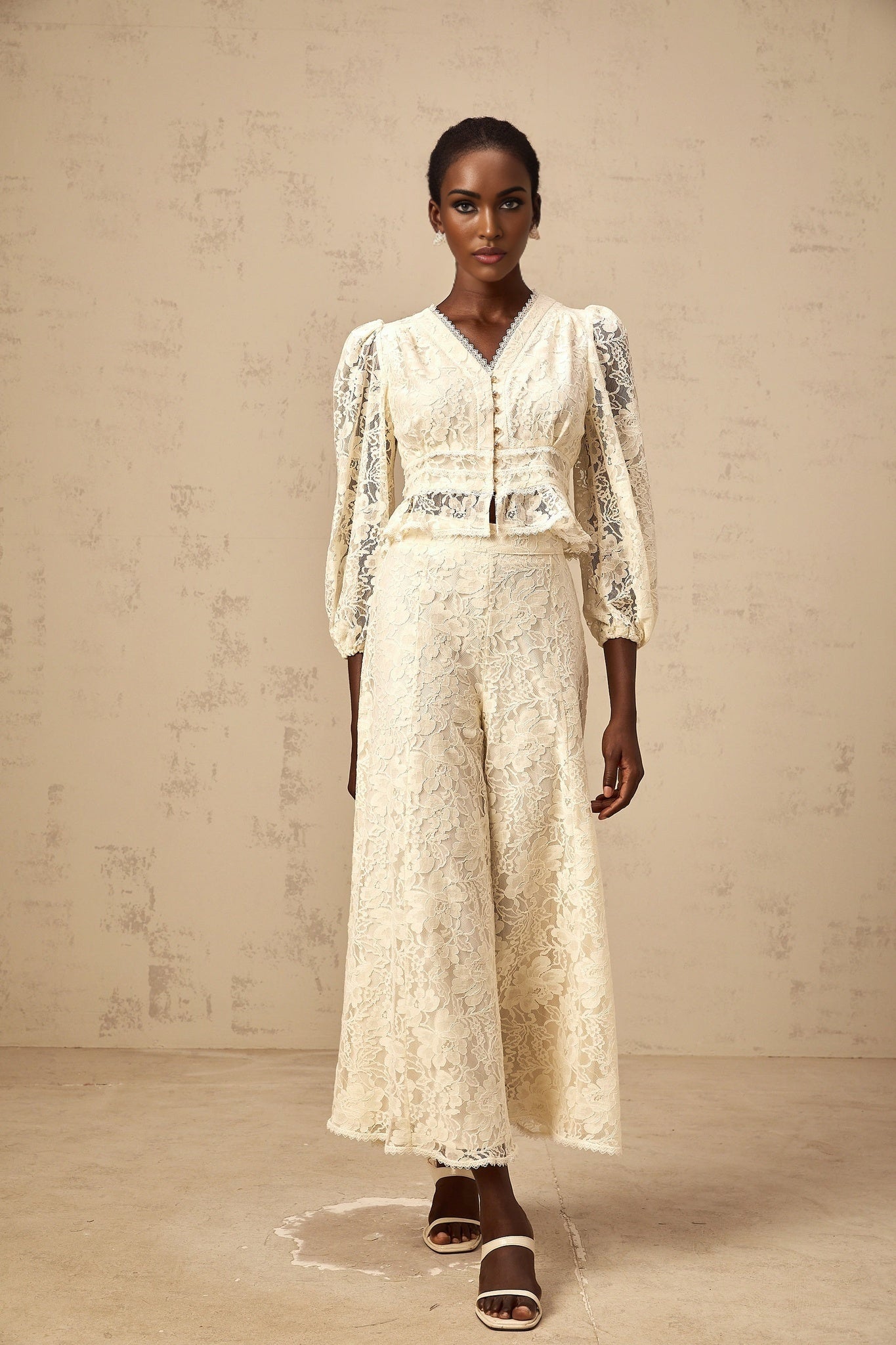 Woman wearing matching beige floral lace blouse and trousers set standing against plain background
