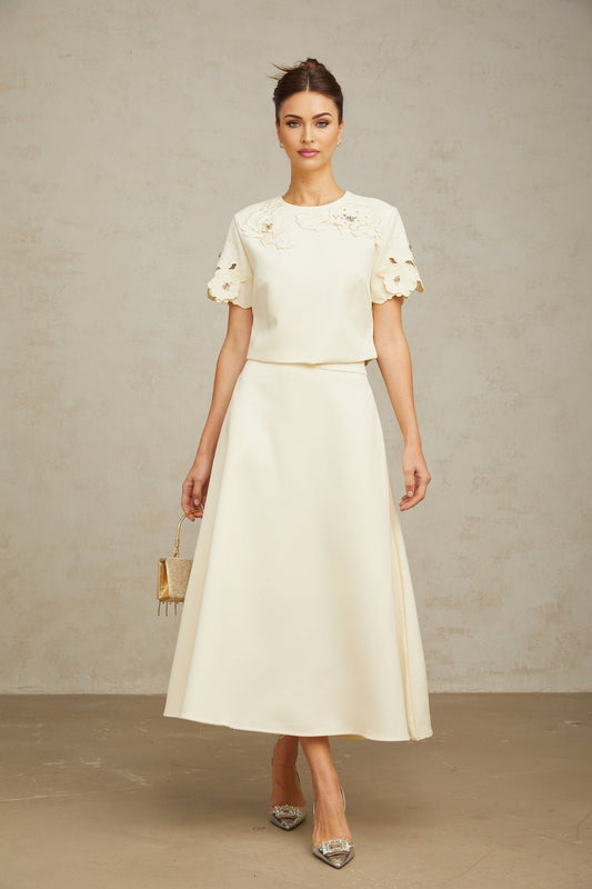 Woman wearing matching neutral color top and skirt set with 3D floral petal details Top has short sleeves and skirt is knee length