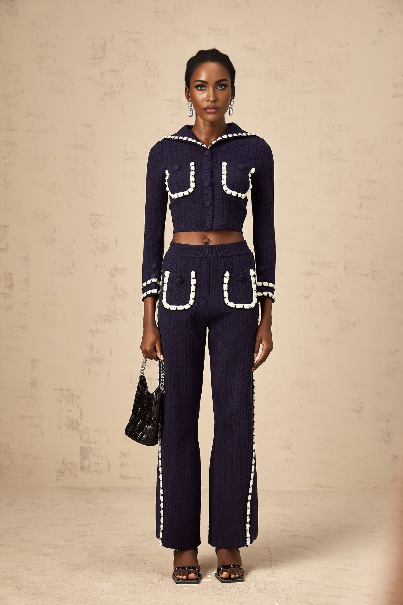 Woman wearing navy blue crochet knit cardigan and matching trousers set standing against plain background