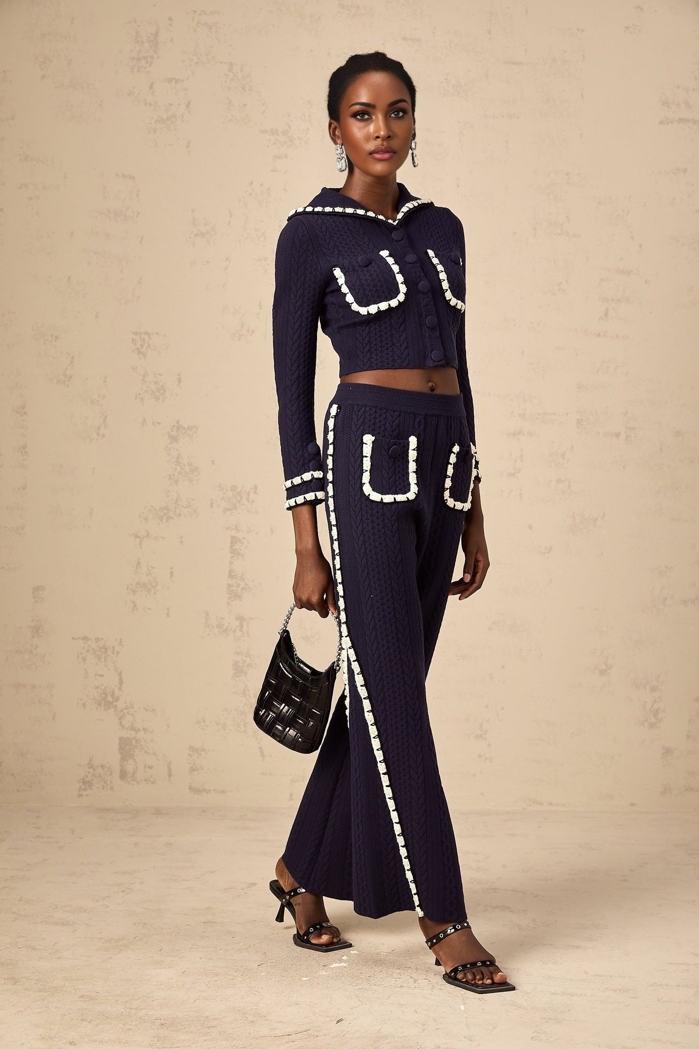 Woman wearing navy blue crochet knit cardigan and matching trousers standing against plain background