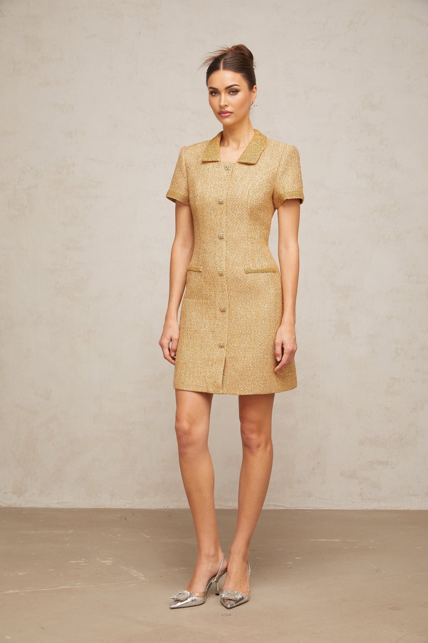 Woman wearing short gold sequined tweed dress with long sleeves and high neckline standing against plain background