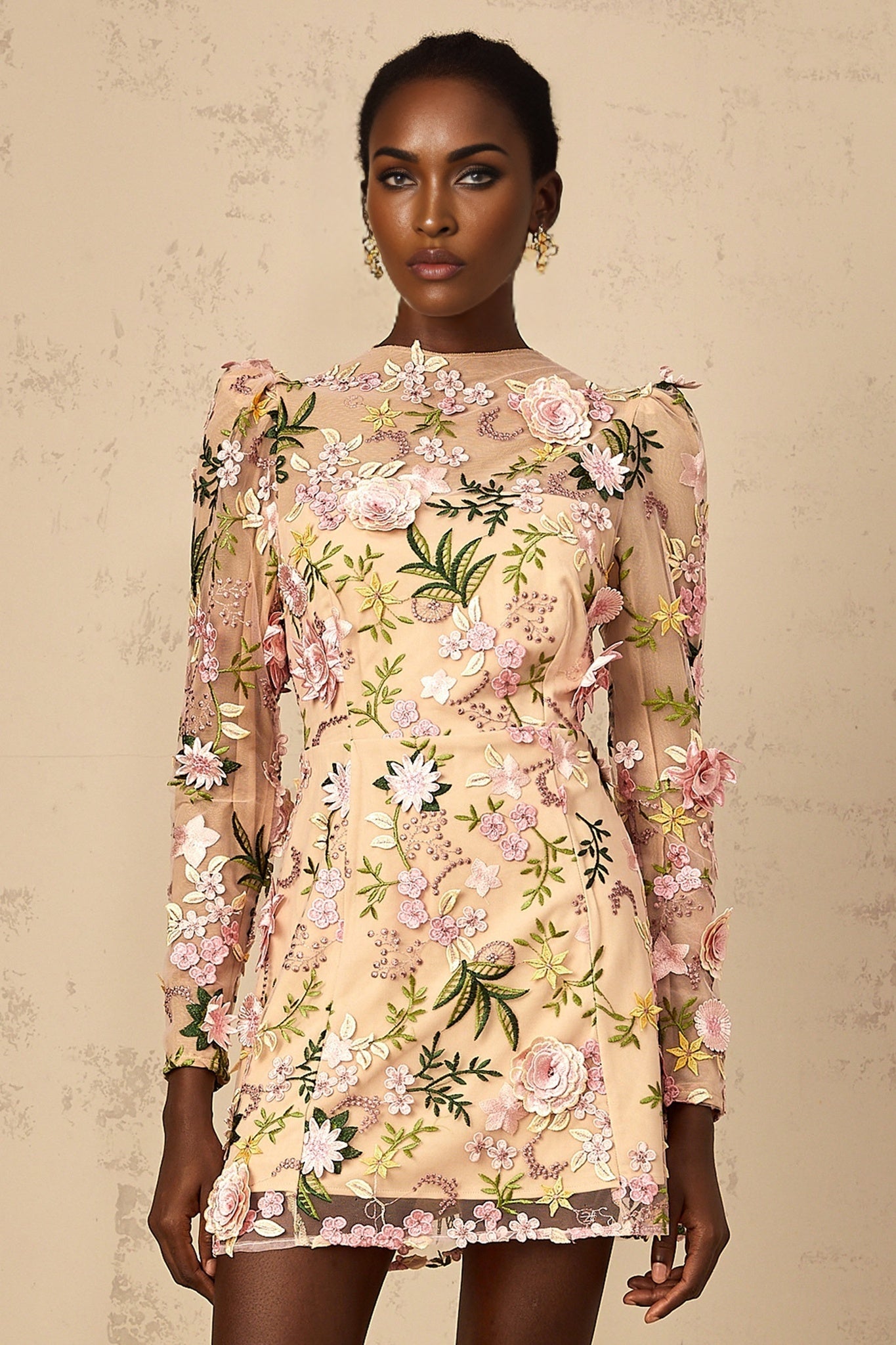 Woman wearing short pink dress with 3D floral applique details standing against plain background