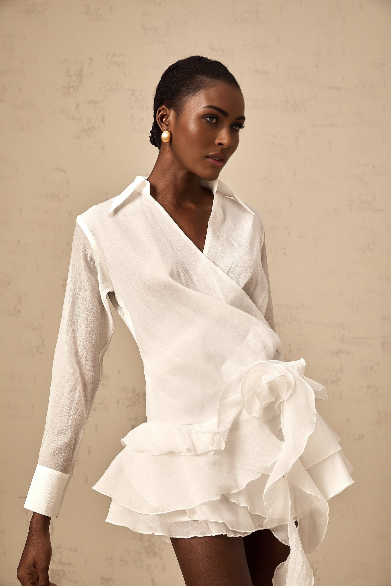 Woman wearing short white semi sheer dress with floral pattern and ruffled details standing against plain background