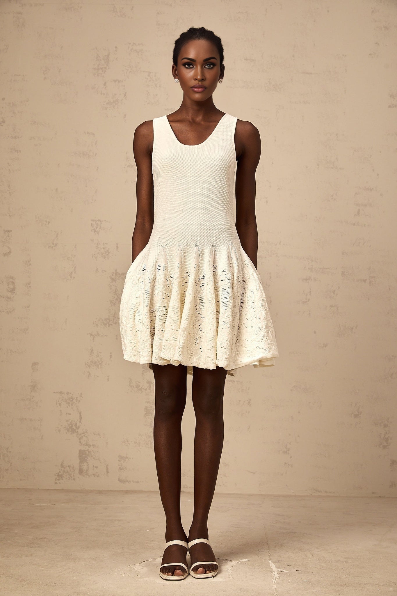 Woman wearing short white textured knit dress with intricate pattern standing against plain background
