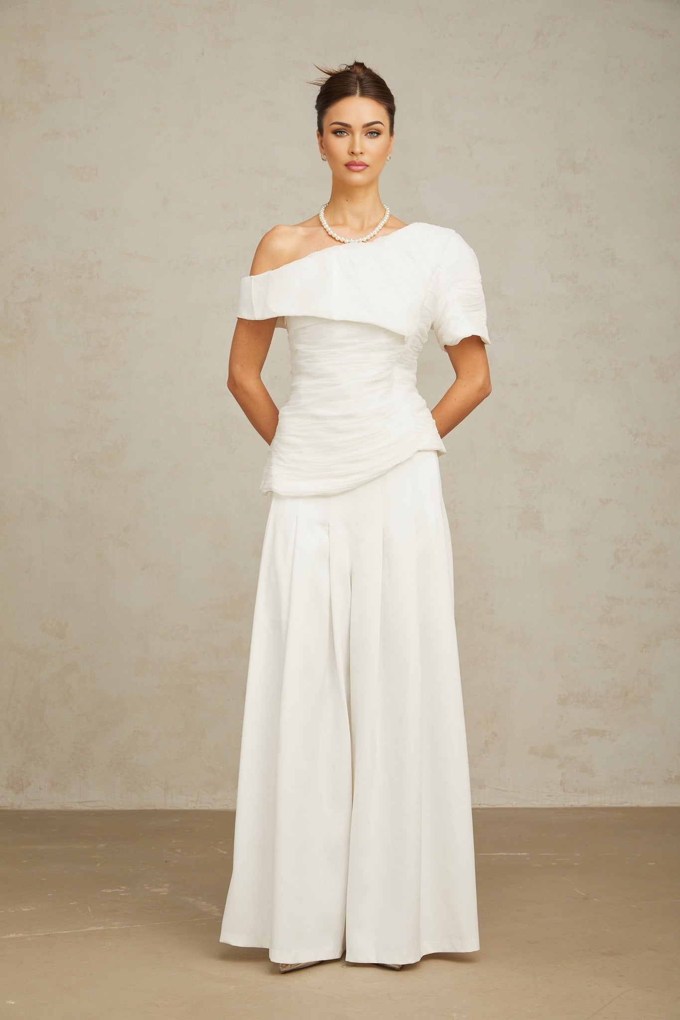 Woman wearing white creased top and matching trousers standing against plain background