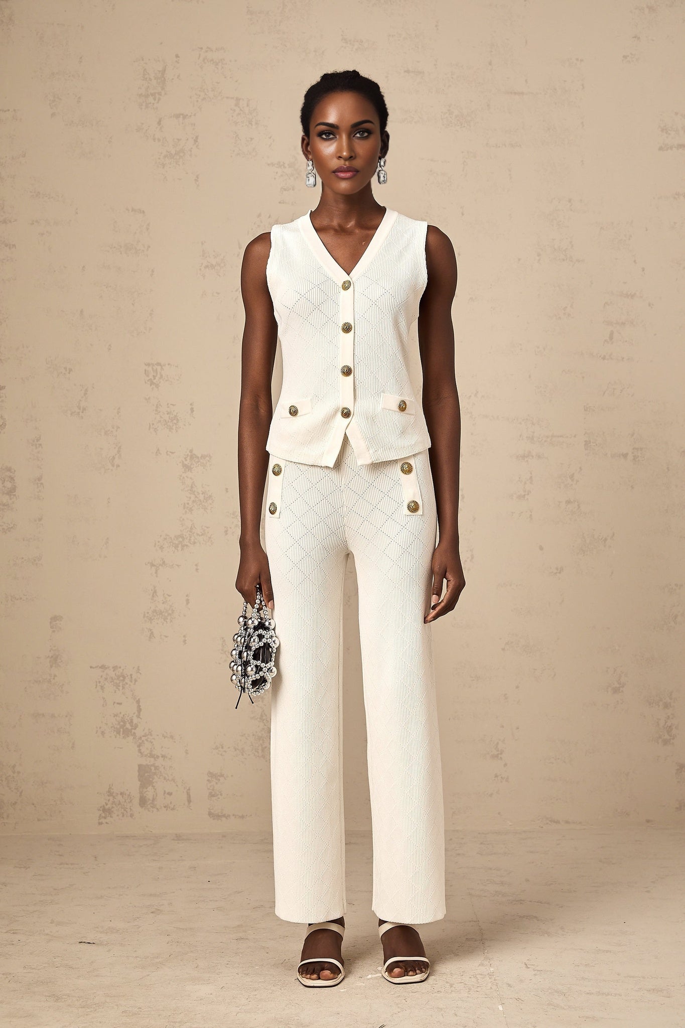 Woman wearing white knitted sleeveless top and matching trousers standing against plain background